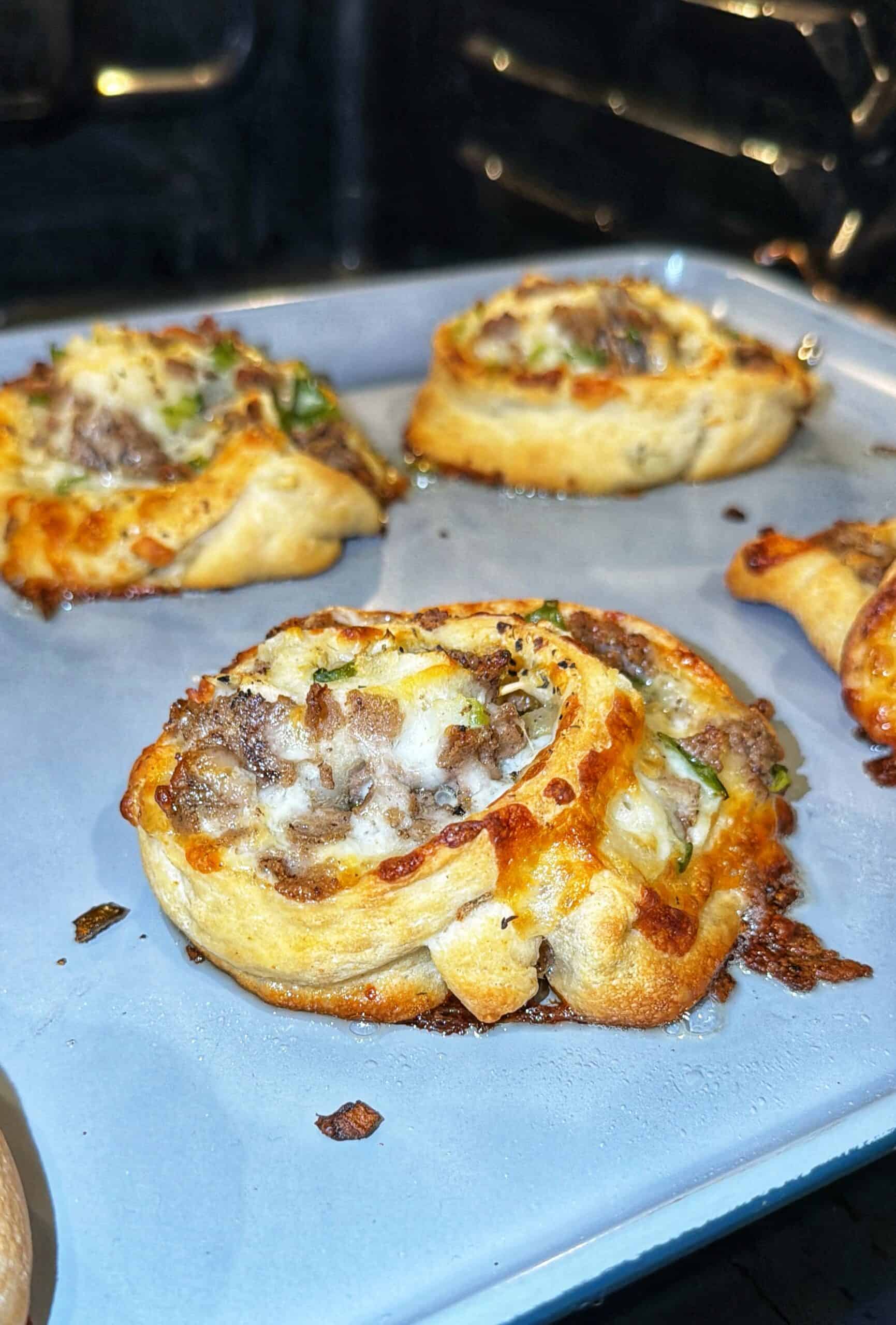 Pizza rolls with melted cheese, ground beef, and herbs baking on a metal tray in an oven. The rolls are golden brown with bubbly cheese and slightly crispy edges.