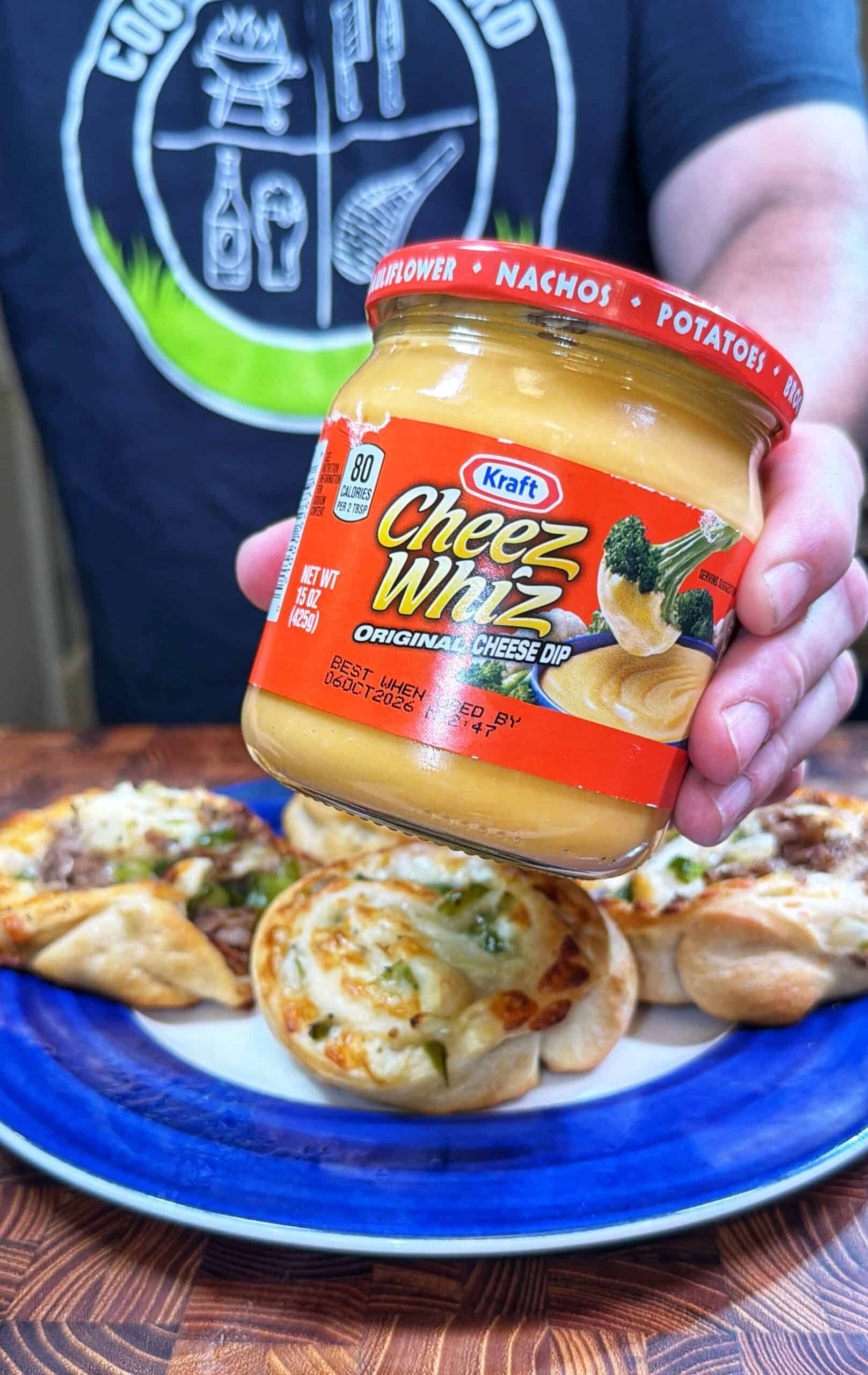 A person holds a jar of Kraft Cheez Whiz original cheese dip above a plate with several baked savory pastries. The person is wearing a dark shirt with a circular graphic.