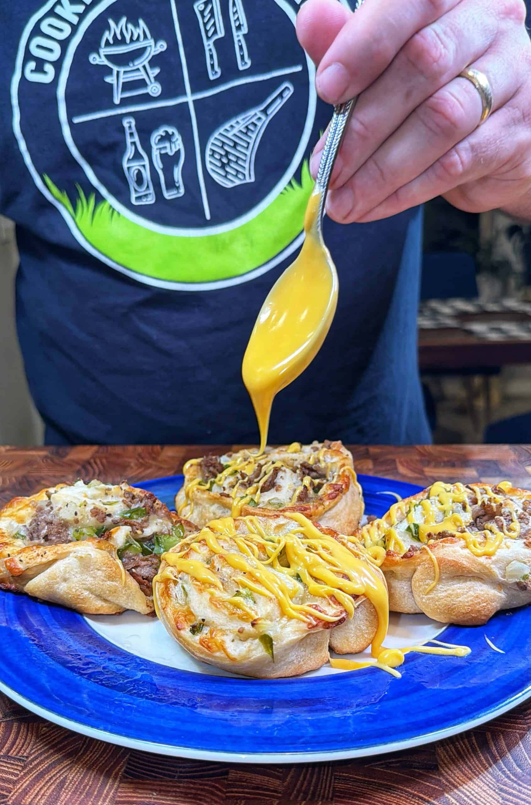 A person drizzles melted cheese over small pizza rolls on a blue plate. The person wears a dark T-shirt with grilling and beverage icons on it. The pizza rolls are topped with various ingredients.