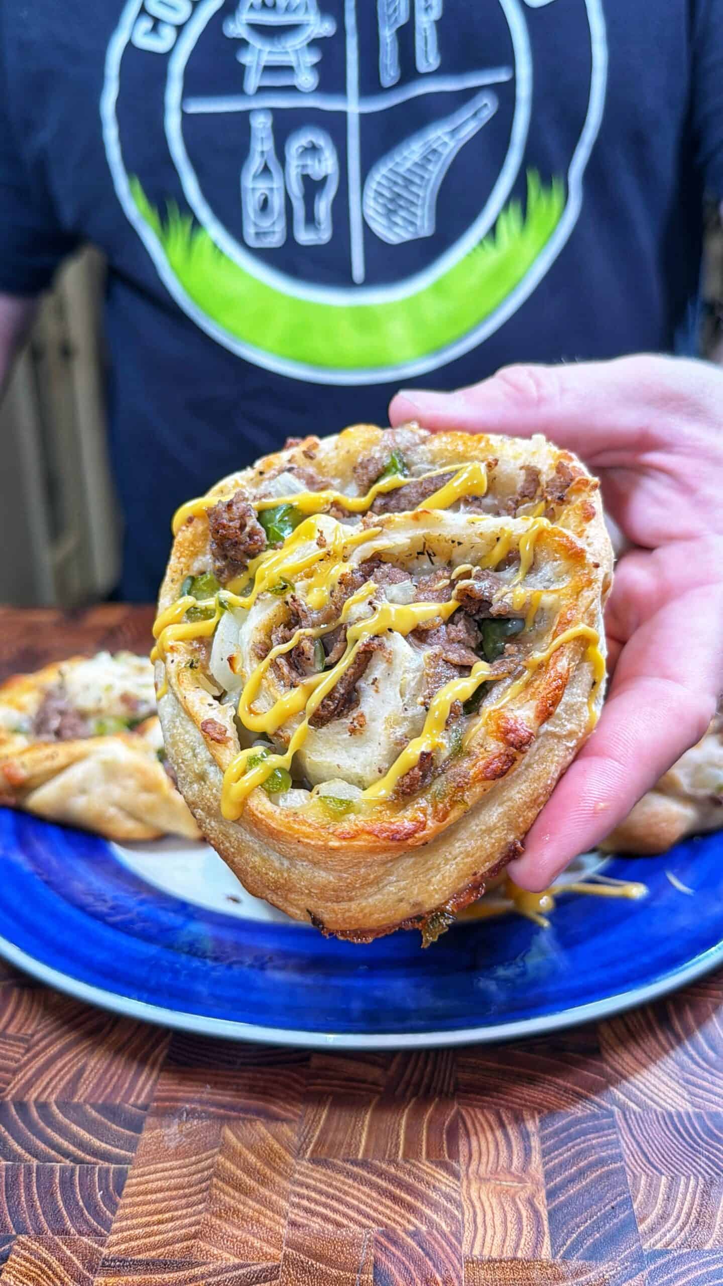 A person holding a savory roll topped with beef, melted cheese, and a drizzle of mustard, over a blue plate. The person is wearing a black shirt with white graphics.