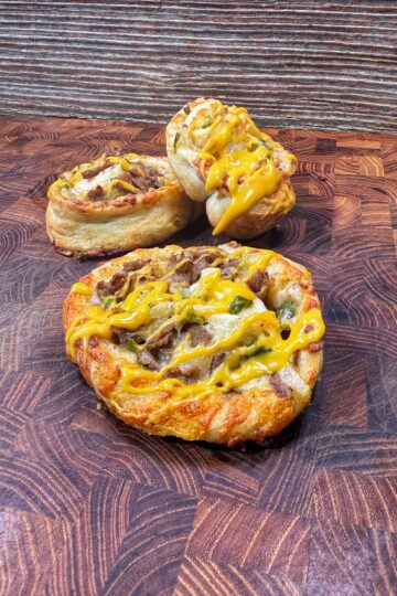 Three cheesesteak pinwheels topped with melted cheese are displayed on a patterned wooden cutting board, with a rustic wooden background behind them.