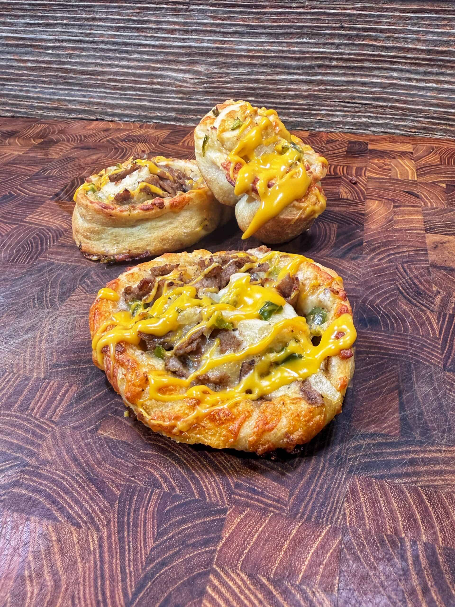 Three cheesesteak pinwheels topped with melted cheese are displayed on a patterned wooden cutting board, with a rustic wooden background behind them.