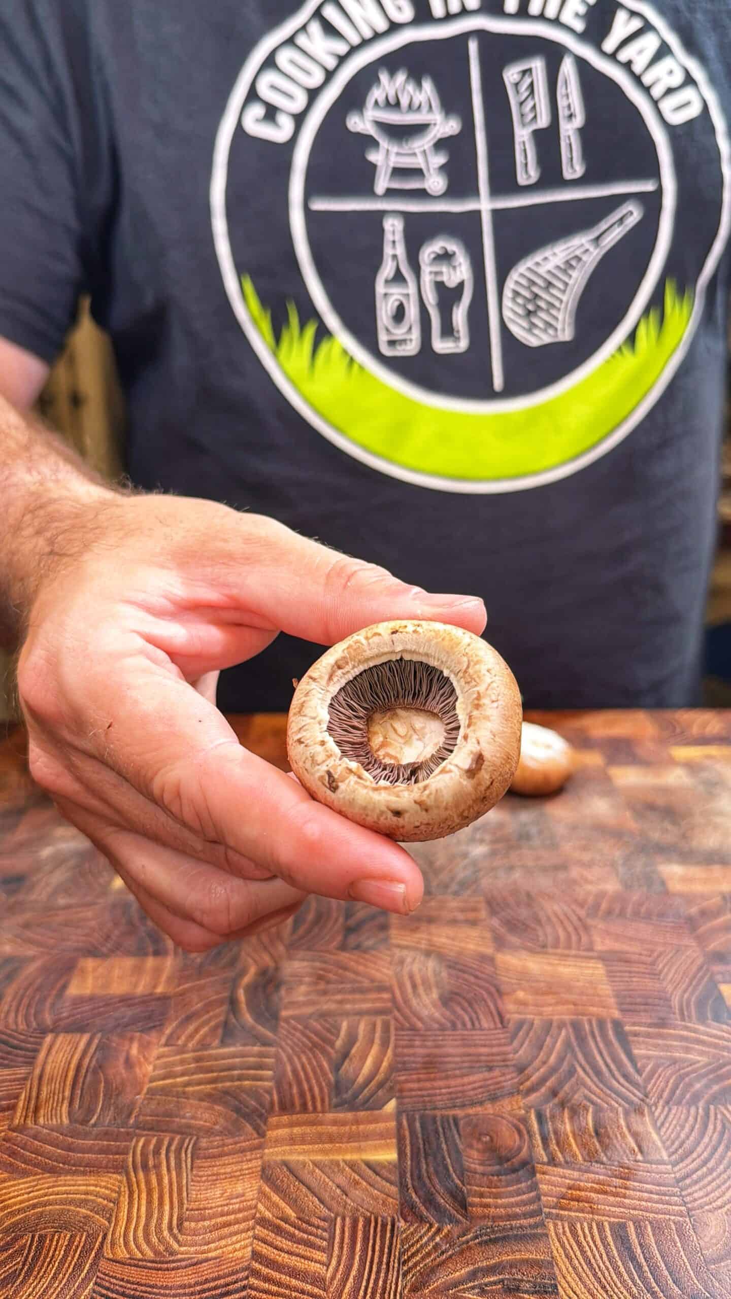 A person wearing a Cooking in the Yard shirt holds a brown mushroom with visible gills over a wooden cutting board.