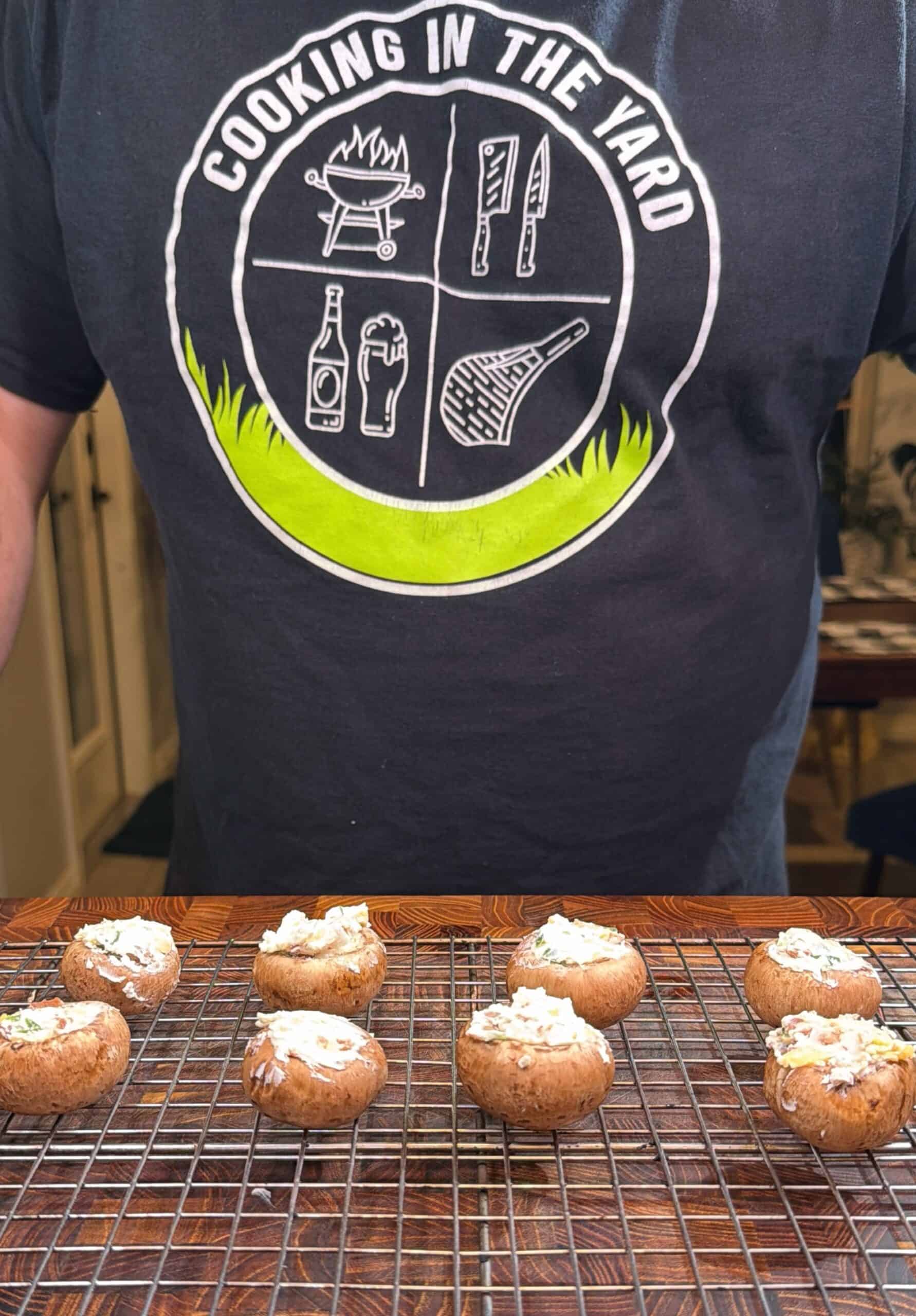 A person wearing a Cooking in the Yard shirt stands behind a cooling rack with stuffed mushrooms arranged in two rows.