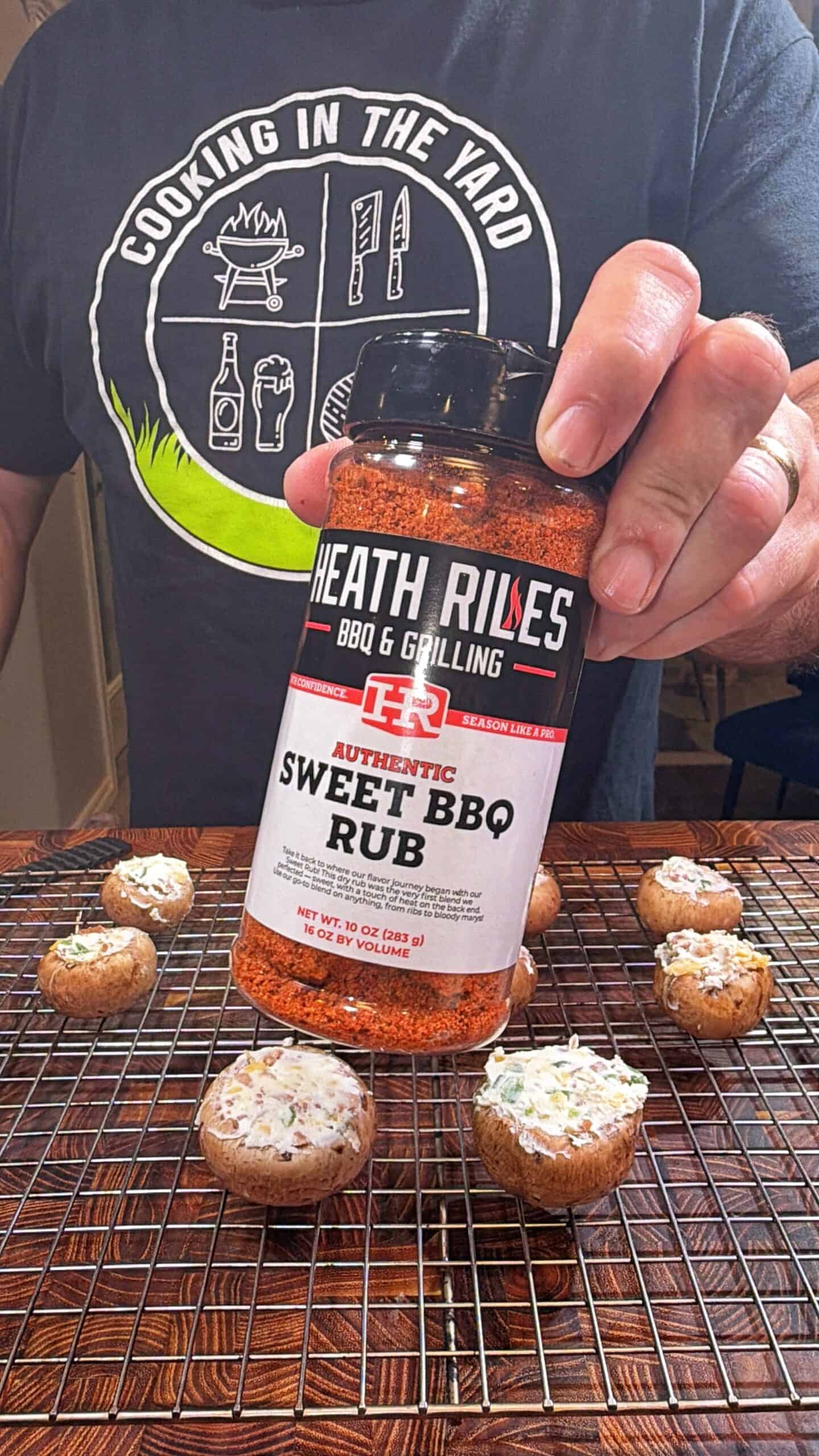 A person wearing a Cooking in the Yard shirt holds a bottle of Heath Riles Authentic Sweet BBQ Rub over a wire rack with stuffed mushrooms, ready for seasoning.