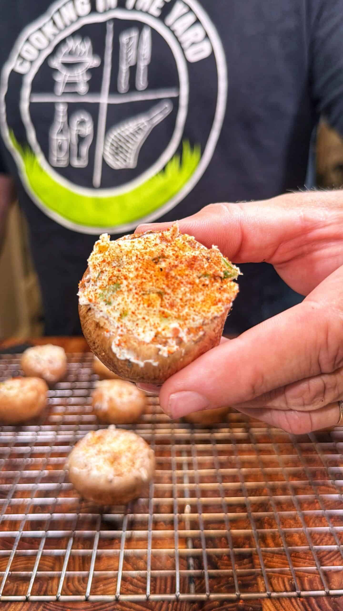 A person holds a stuffed mushroom topped with seasoning. More stuffed mushrooms are on a wire rack below. The person is wearing a dark shirt with a green and white logo.