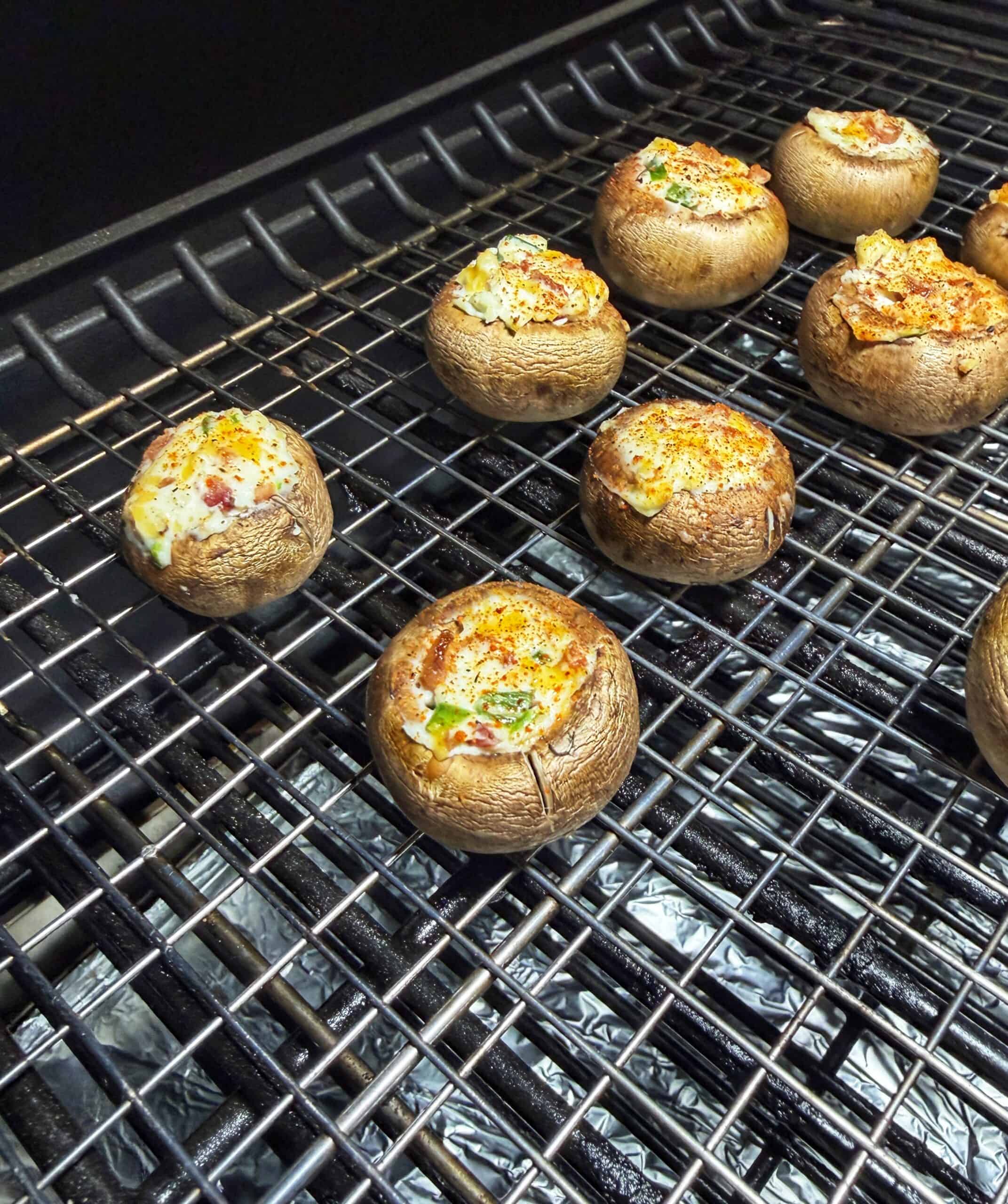 Stuffed mushrooms on a grill, filled with a creamy mixture and sprinkled with herbs and seasoning, are being cooked; the mushrooms are arranged across the metal grates.