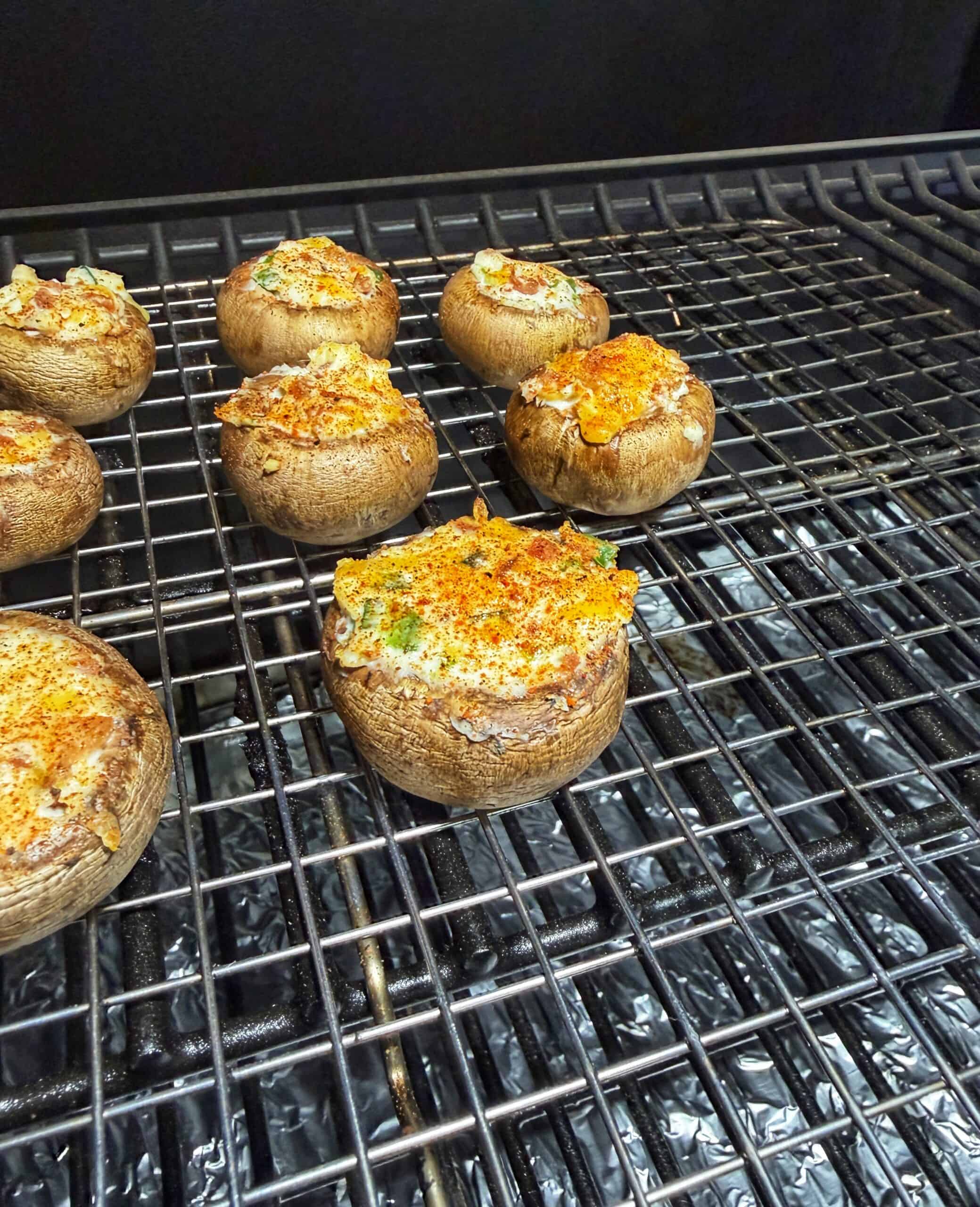 Stuffed mushrooms topped with herbs and spices are arranged on a grill rack, ready to be cooked. The mushrooms appear filled with a creamy mixture and are spaced apart on the metal grill grate.