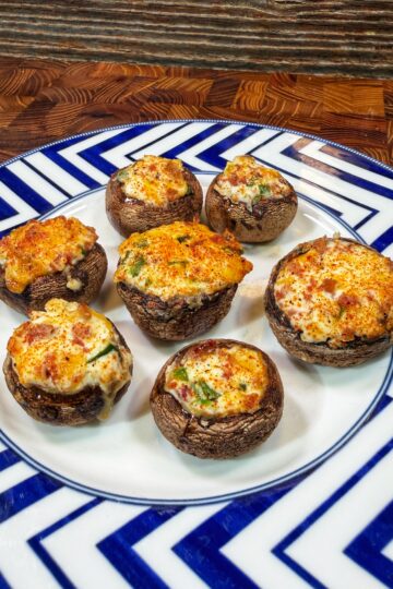 A plate with seven stuffed mushrooms, topped with a golden, cheesy filling, sits on a geometric blue-and-white patterned plate on a wooden surface.