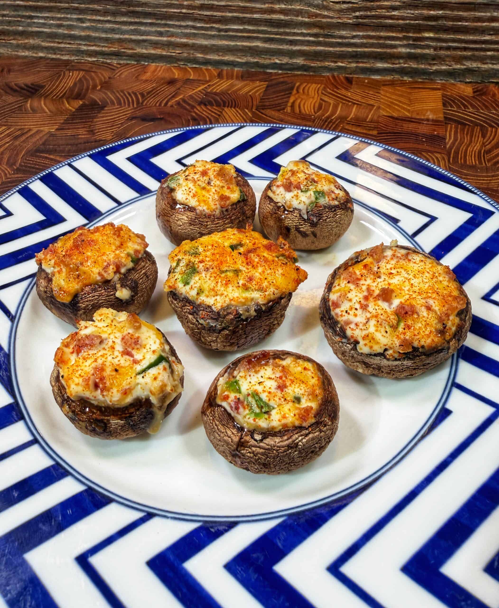A plate with seven stuffed mushrooms, topped with a golden, cheesy filling, sits on a geometric blue-and-white patterned plate on a wooden surface.