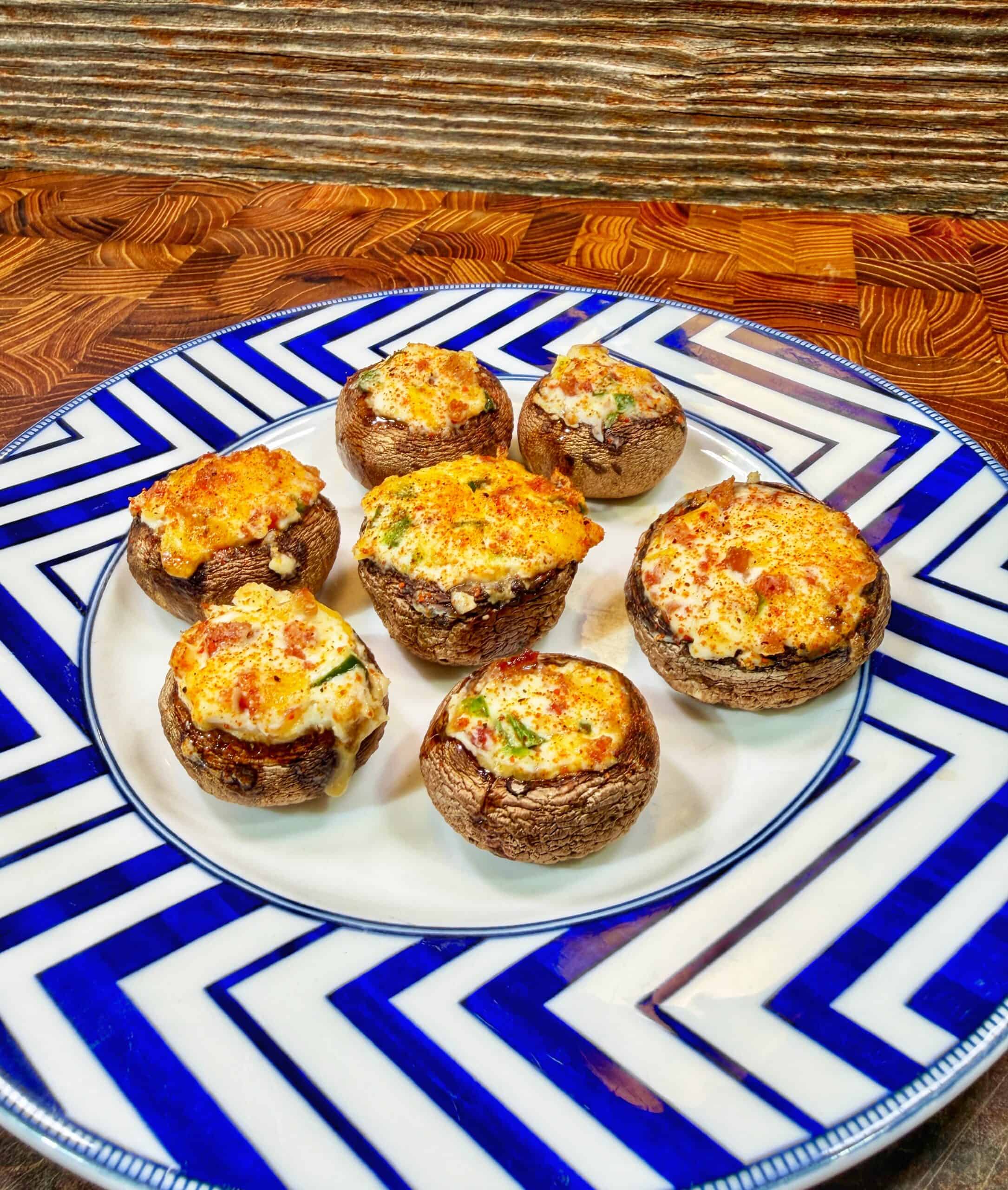 A plate with seven stuffed mushrooms topped with melted cheese and herbs, arranged on a white dish with a bold blue zigzag border, set on a wooden surface.