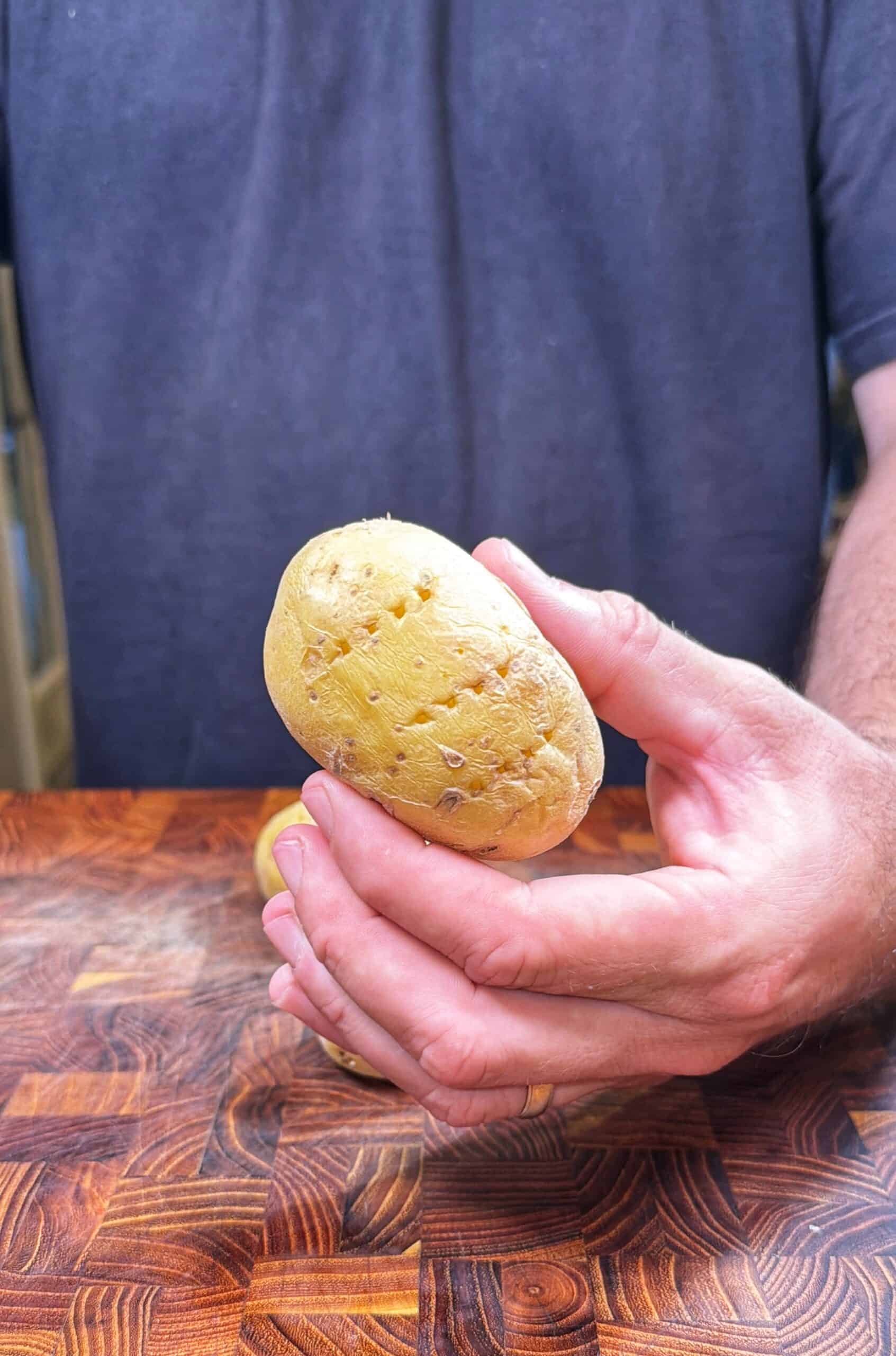A person wearing a dark shirt holds a potato with small holes in it, standing behind a wooden surface.