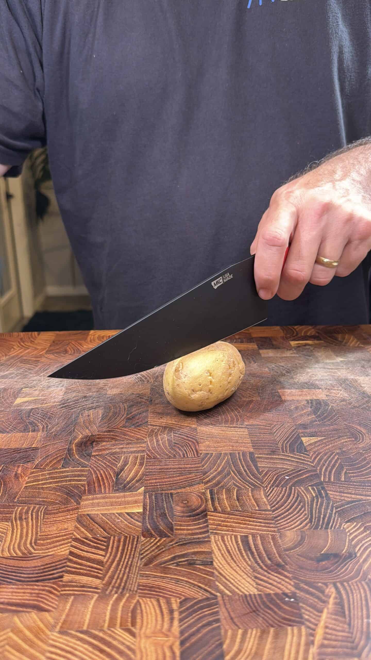A person holds a black knife above a small potato on a wooden cutting board, preparing to slice it. Only the persons torso and hand are visible.