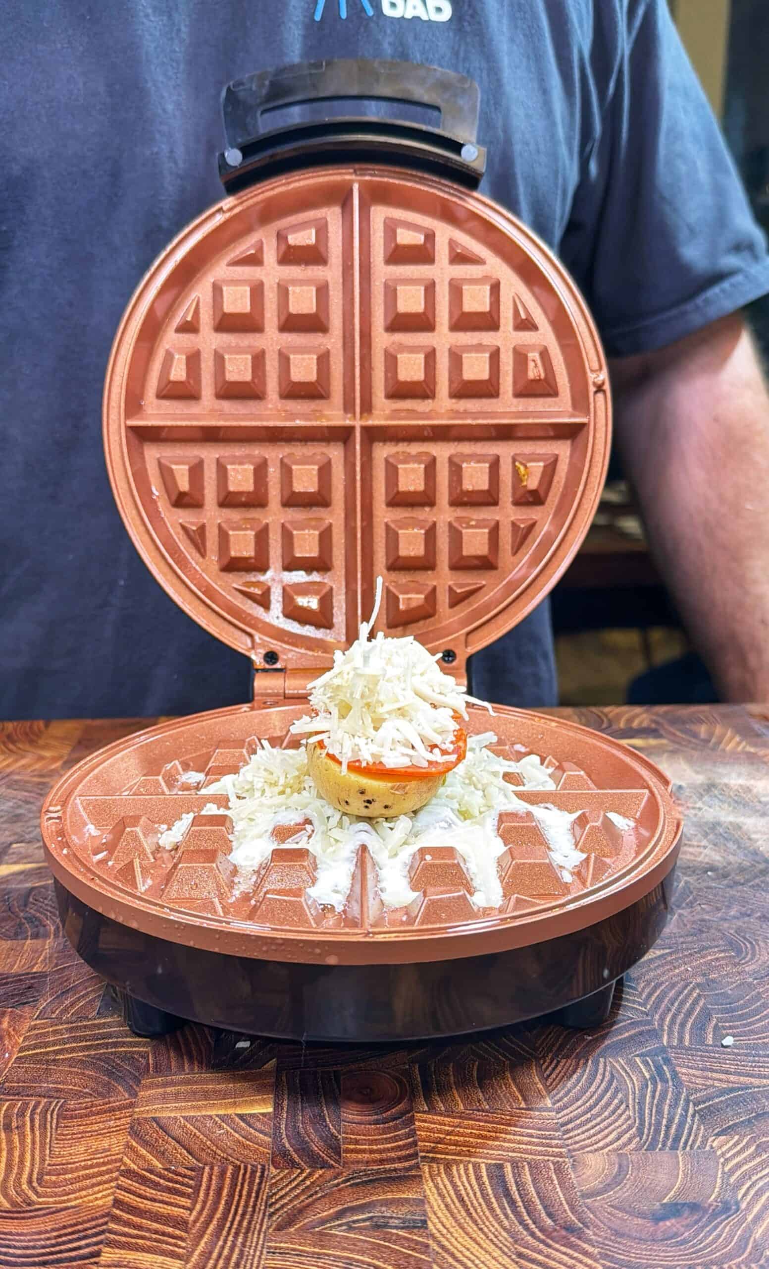 A person stands behind an open waffle maker on a wooden counter. Inside, a raw egg, sliced tomato, and shredded cheese sit on the lower plate, ready to be cooked.