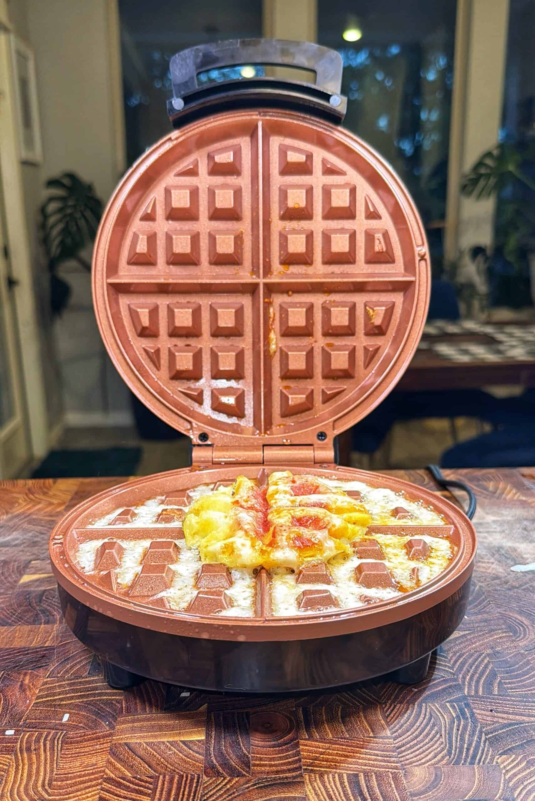 A waffle maker sits open on a wooden counter, with melted cheese and sauce oozing out from a messy waffle inside. The background shows a window and some indoor plants.
