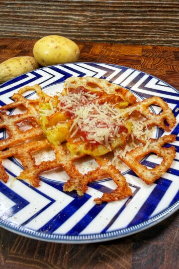 A cheesy potato waffle topped with shredded cheese sits on a blue and white patterned plate. Two whole potatoes are in the background on a wooden surface.