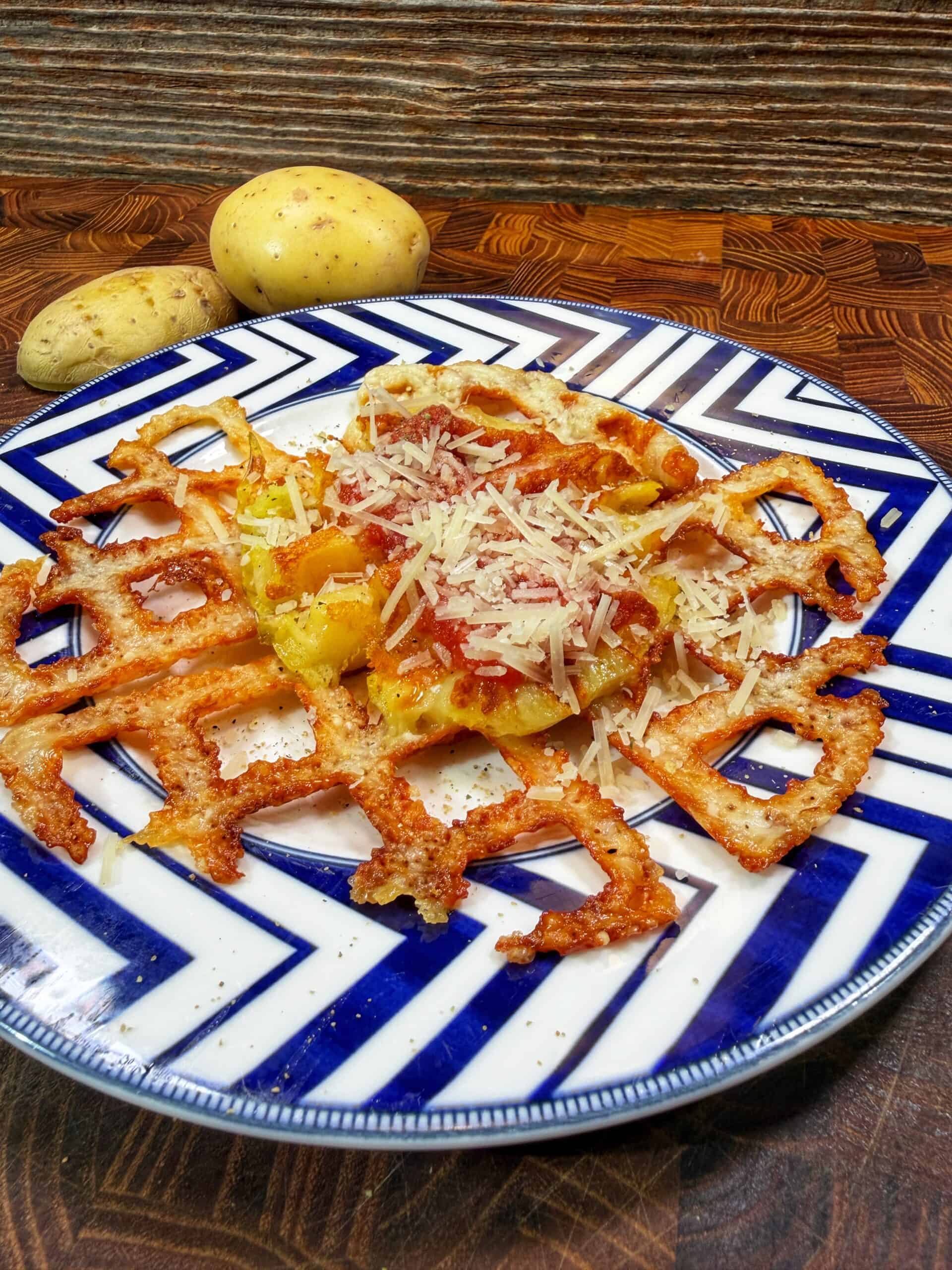 A cheesy potato waffle topped with shredded cheese sits on a blue and white patterned plate. Two whole potatoes are in the background on a wooden surface.