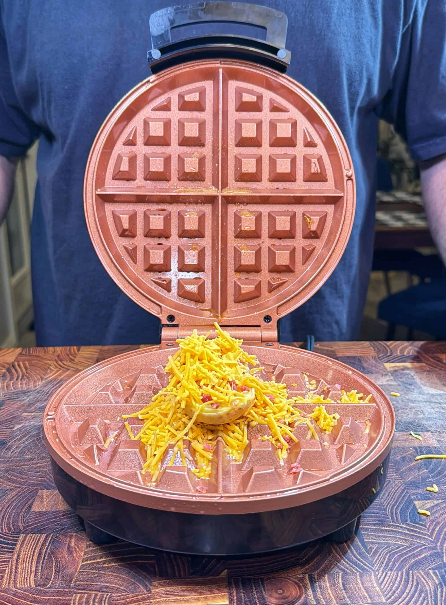 A waffle maker sits open on a wooden counter with a pile of shredded cheddar cheese in the center, ready to be cooked. A person in a blue shirt stands behind the waffle maker.