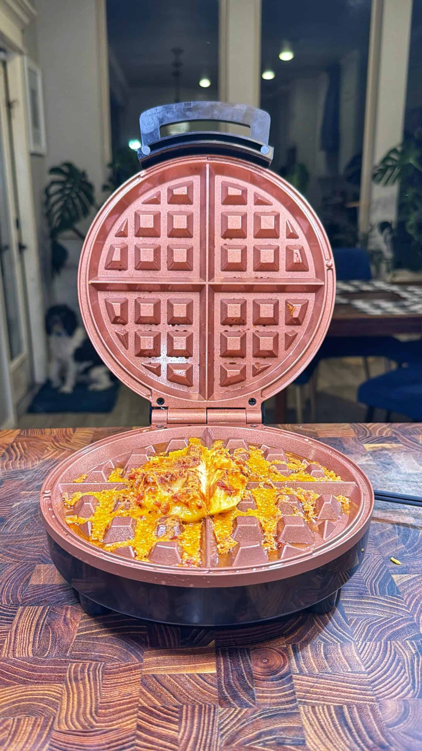 An open waffle maker sits on a wooden counter with a cooked, cheesy food item inside. Cheese is melted and crisp around the edges. A dog is visible in the background near a dining table and plants.