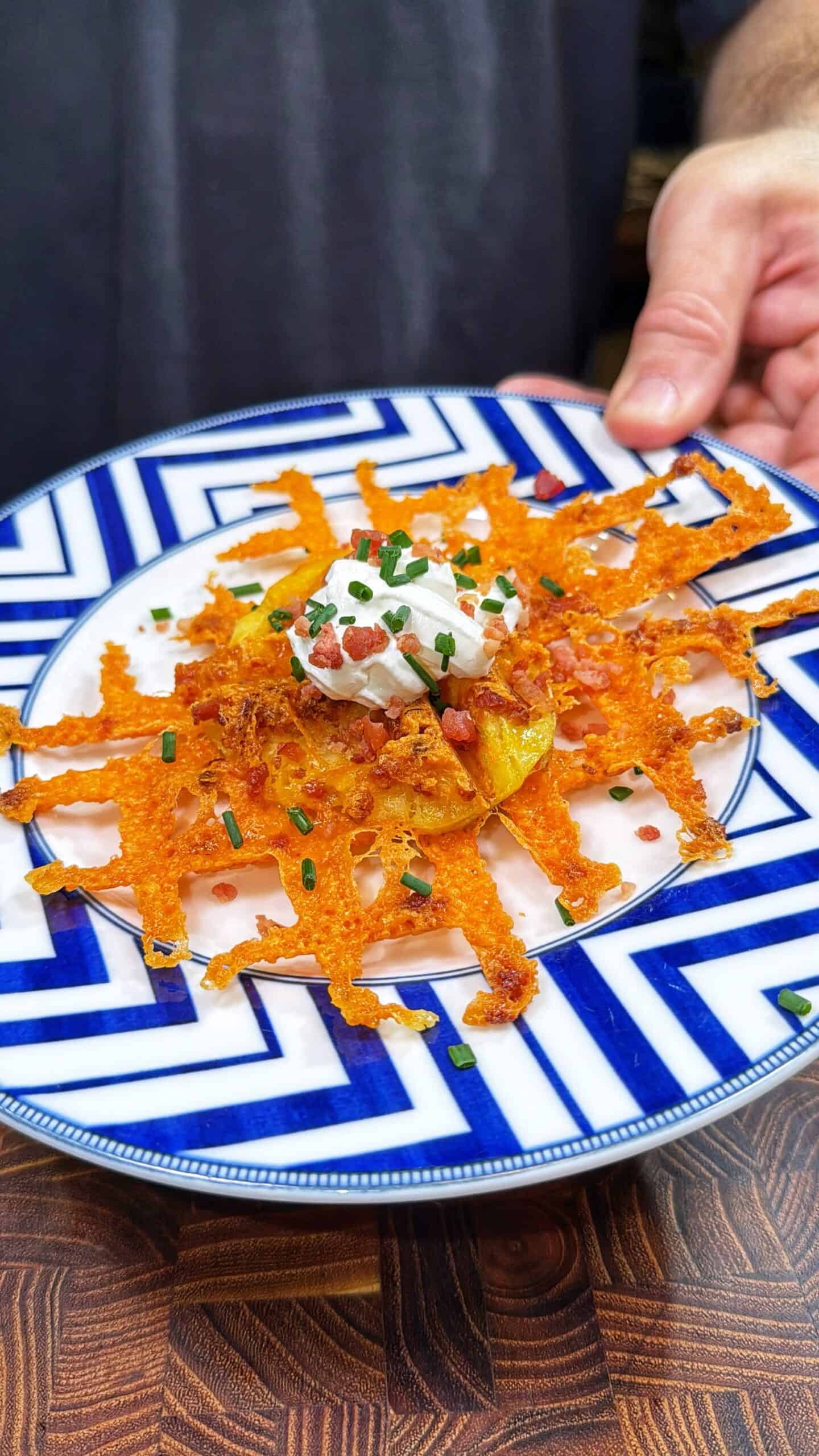 A hand holds a blue and white plate with a crispy, lattice-shaped cheddar cheese nest topped with sour cream, chives, and bacon bits, served on a wooden surface.