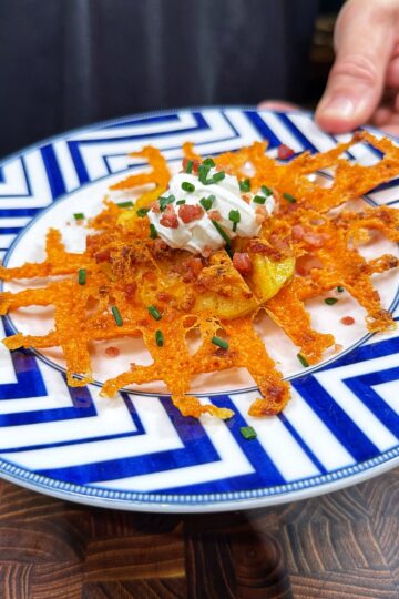 A hand holds a plate with a crispy cheese lattice topped with potato slices, sour cream, bacon bits, and chopped chives, served on a blue and white geometric patterned dish.