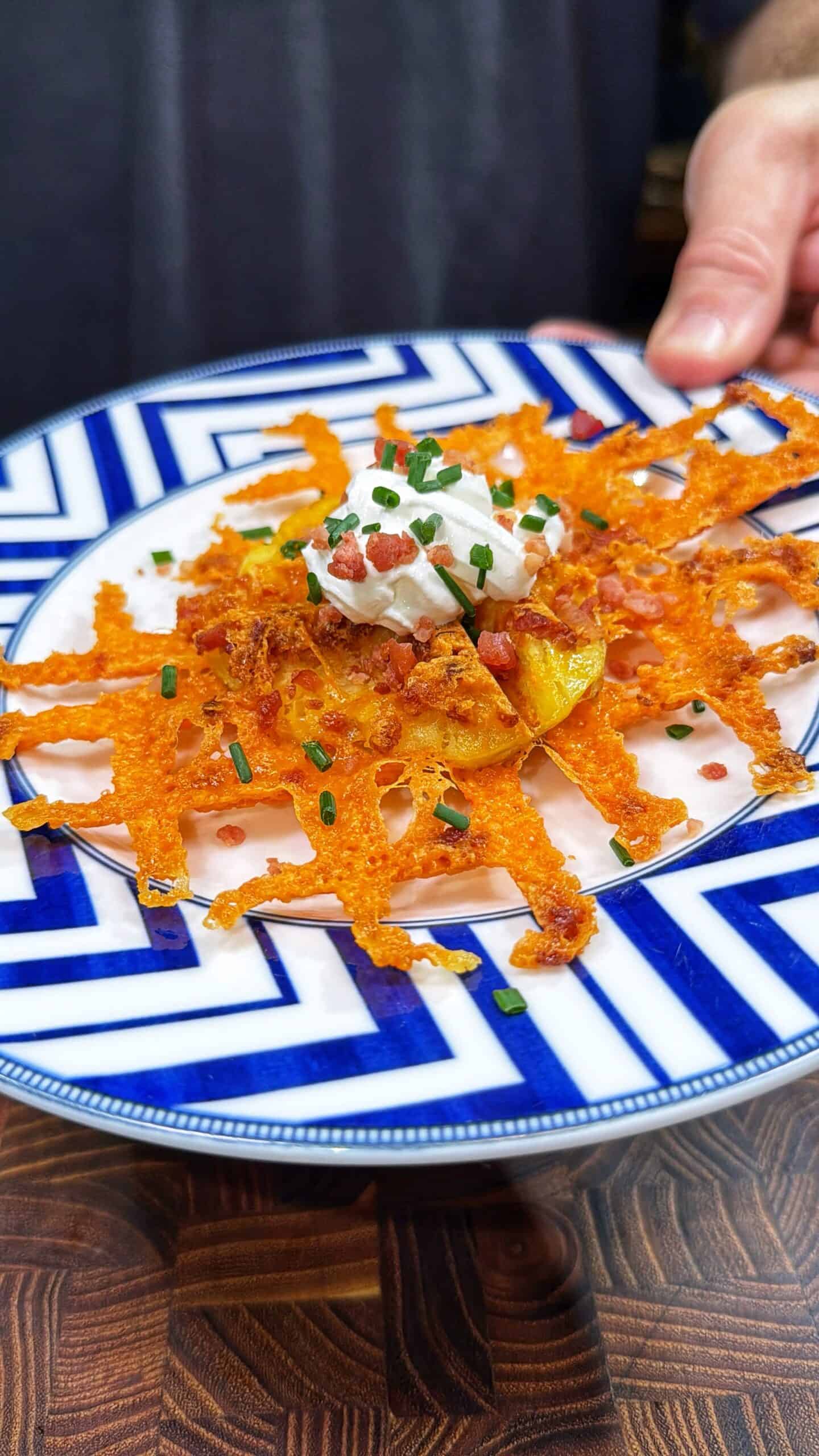 A hand holds a plate with a crispy cheese lattice topped with potato slices, sour cream, bacon bits, and chopped chives, served on a blue and white geometric patterned dish.