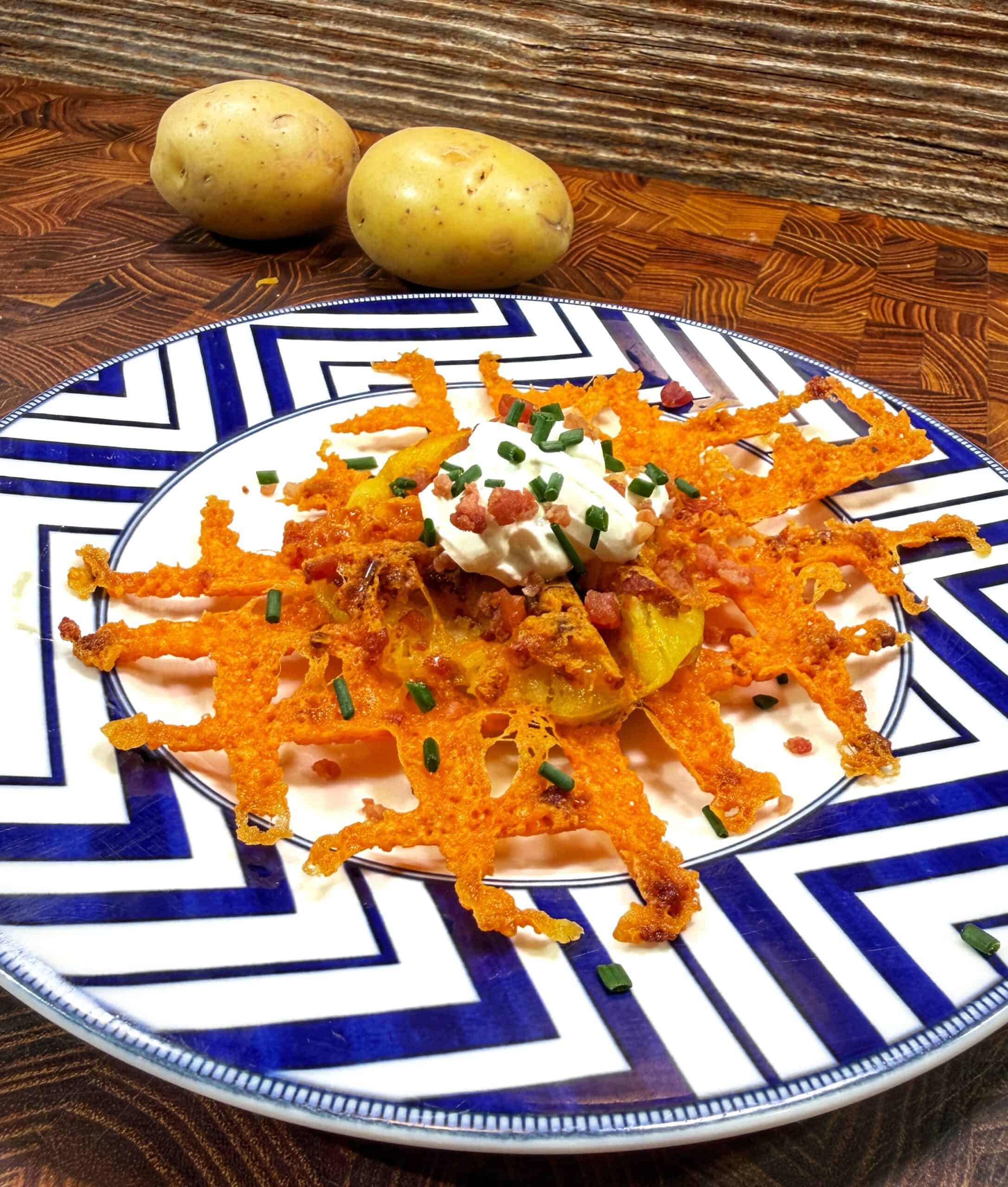 A plate with a crispy cheese and potato dish topped with sour cream, bacon bits, and chopped chives, sitting on a patterned plate. Two whole potatoes are in the background on a wooden surface.