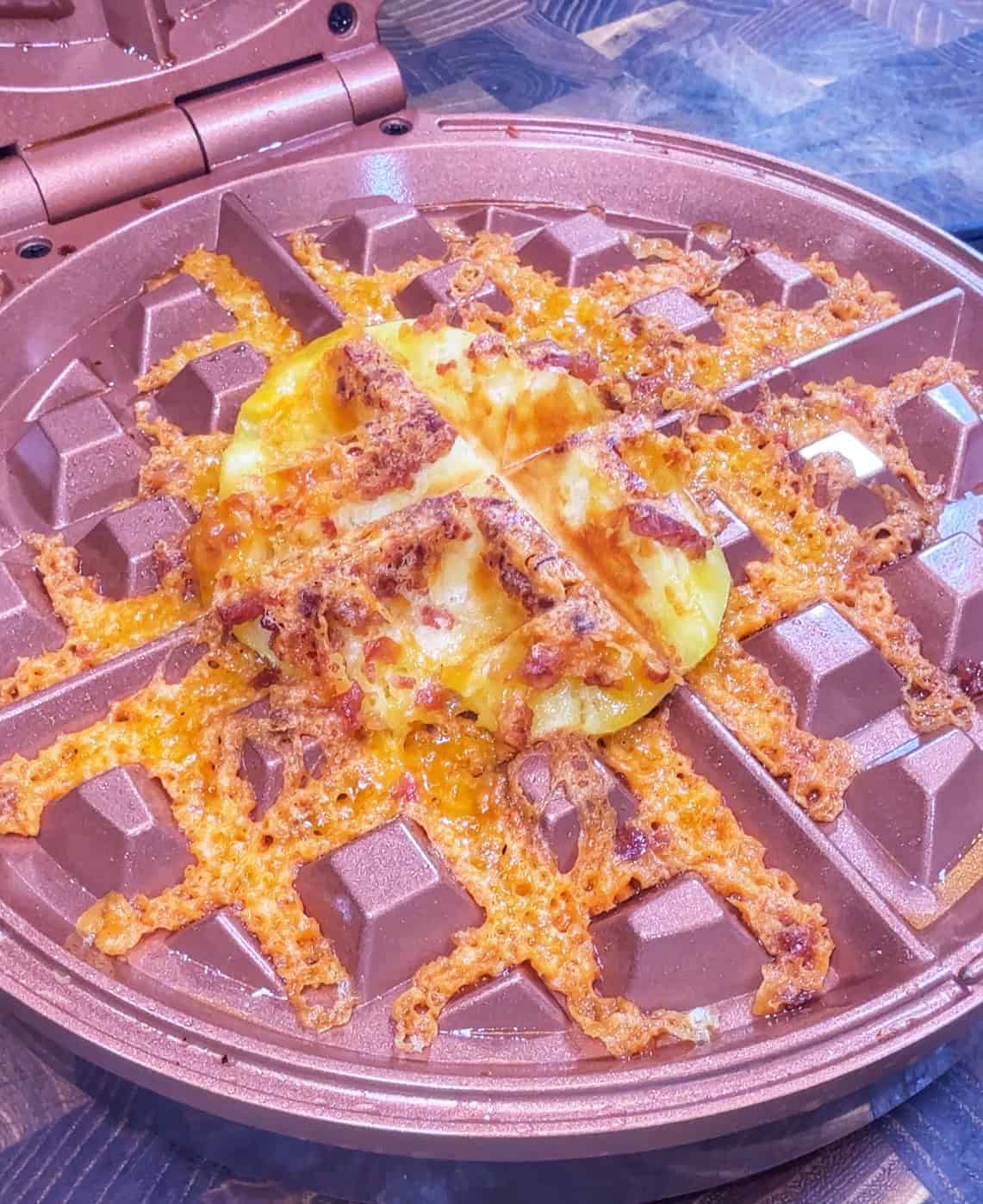 An omelette with crispy edges and melted cheese is cooked in a waffle maker, with cheese and bits spilling over onto the waffle maker’s surface.