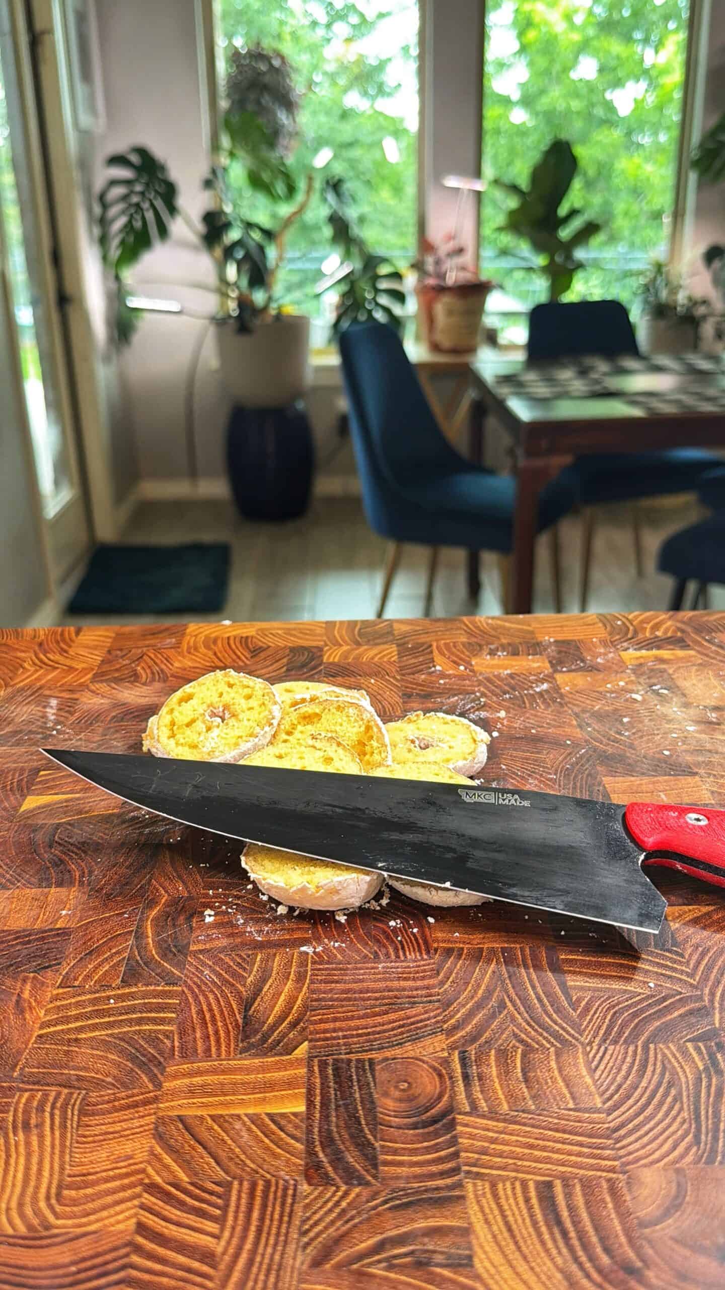 A kitchen knife with a red handle rests on a wooden cutting board, slicing through rounds of bread or pastry. In the background, a dining table, blue chairs, and green plants are visible near large windows.
