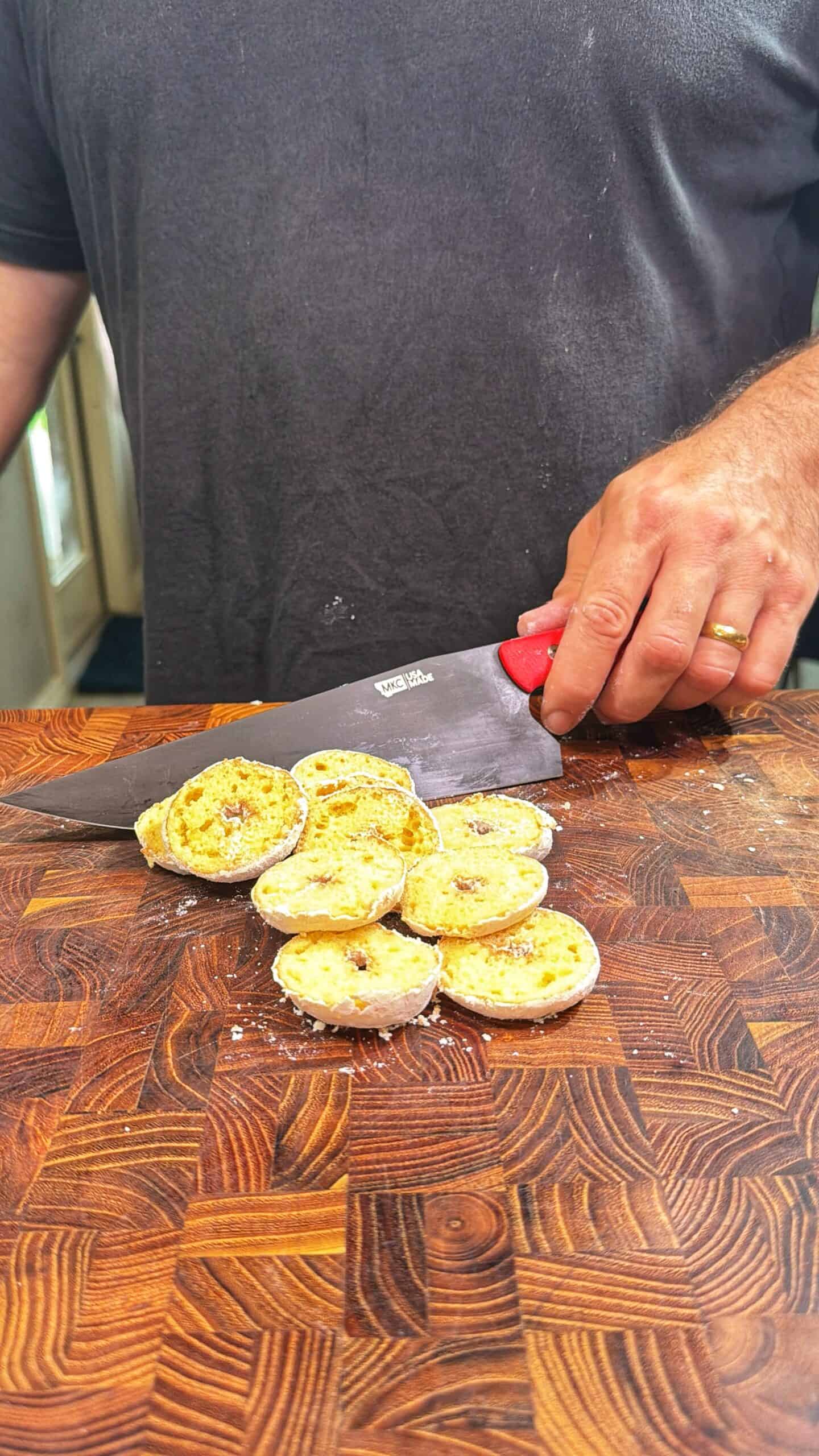 A person slices a banana with a large knife on a patterned wooden cutting board. The banana is cut into round pieces, and the person is wearing a dark shirt and a gold ring on their left hand.