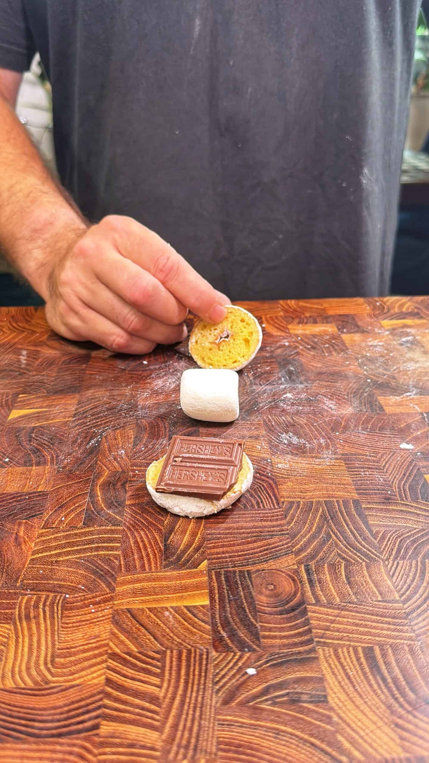 A person assembles a smore on a wooden surface, placing a marshmallow between two graham crackers, with a piece of Hersheys chocolate on one cracker.