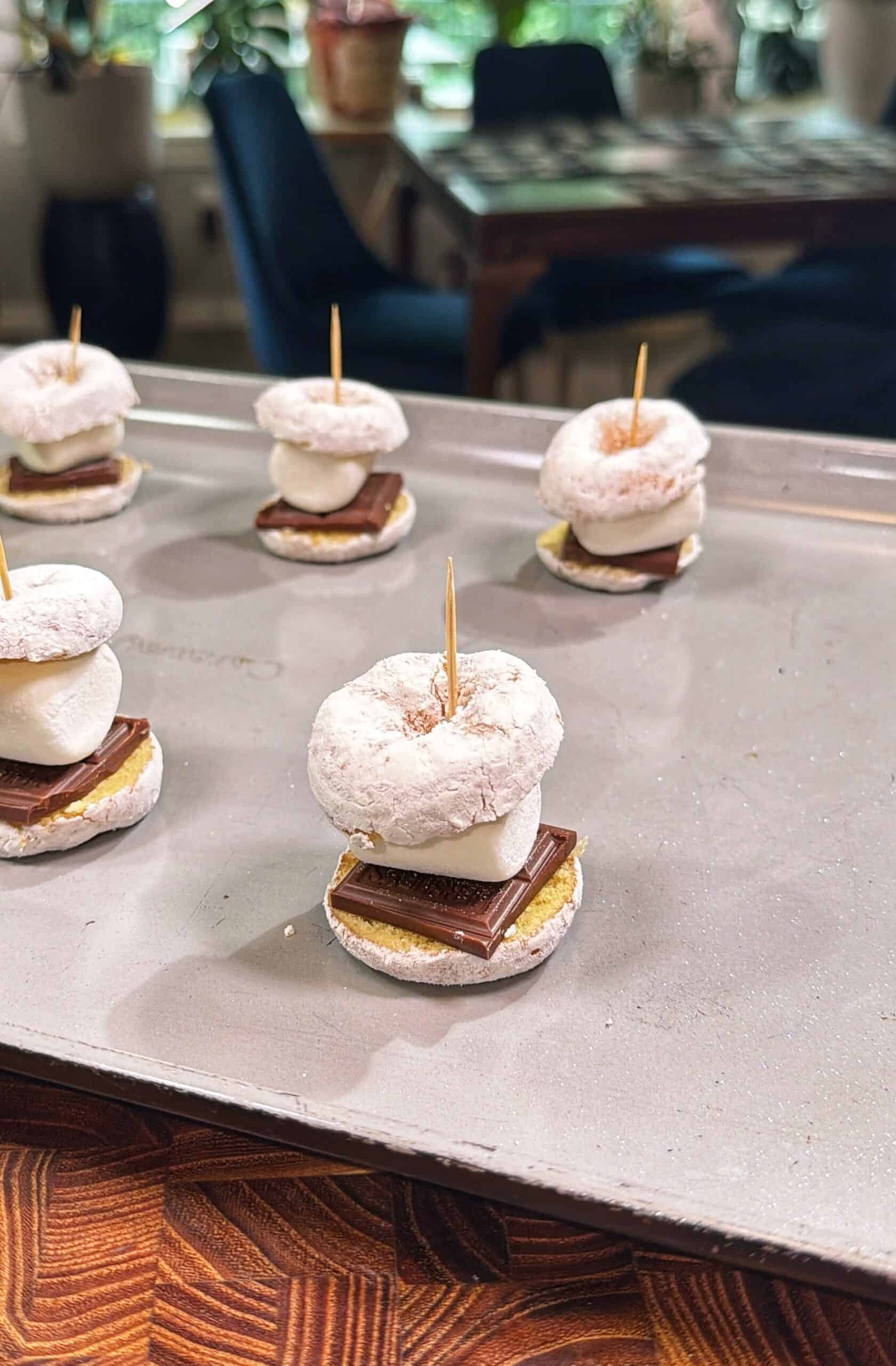 Powdered donuts, marshmallows, chocolate squares, and graham crackers are stacked and held together with toothpicks on a baking sheet, forming a twist on classic smores. A table and chairs are visible in the background.