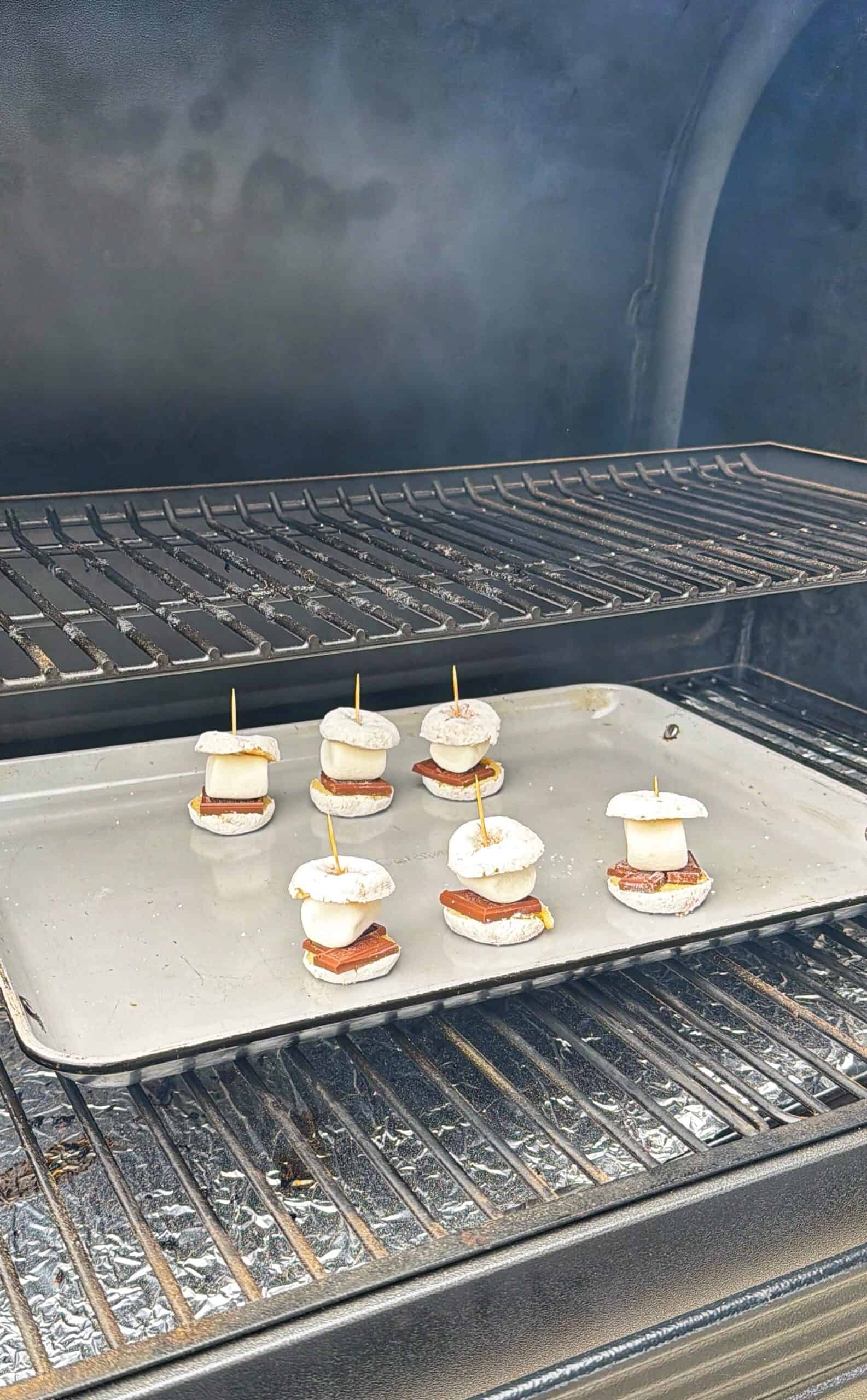 Several small s’mores treats, each held together with a toothpick, sit on a baking sheet inside a smoky grill. Each treat has a marshmallow, chocolate, and cracker layer.