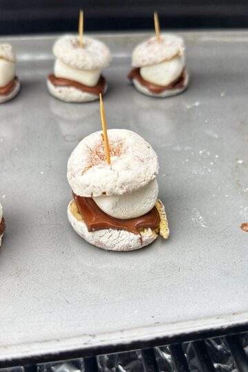 A close-up of a mini s’more made with a powdered donut, chocolate, marshmallow, and graham cracker, held together by a toothpick and placed on a metal baking sheet with more s’mores in the background.