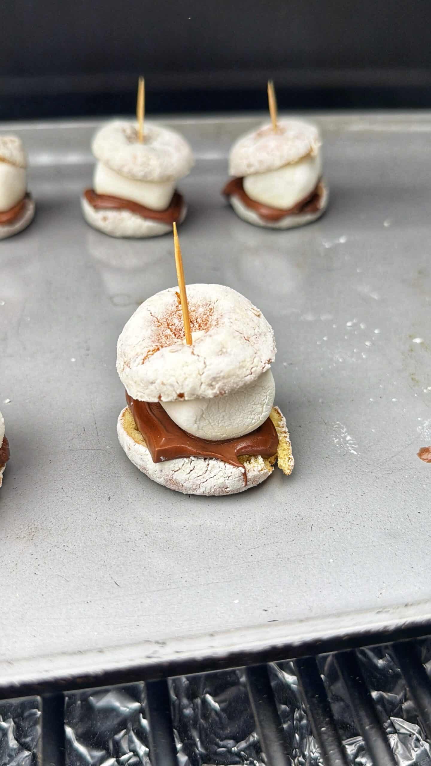 A close-up of a mini s’more made with a powdered donut, chocolate, marshmallow, and graham cracker, held together by a toothpick and placed on a metal baking sheet with more s’mores in the background.