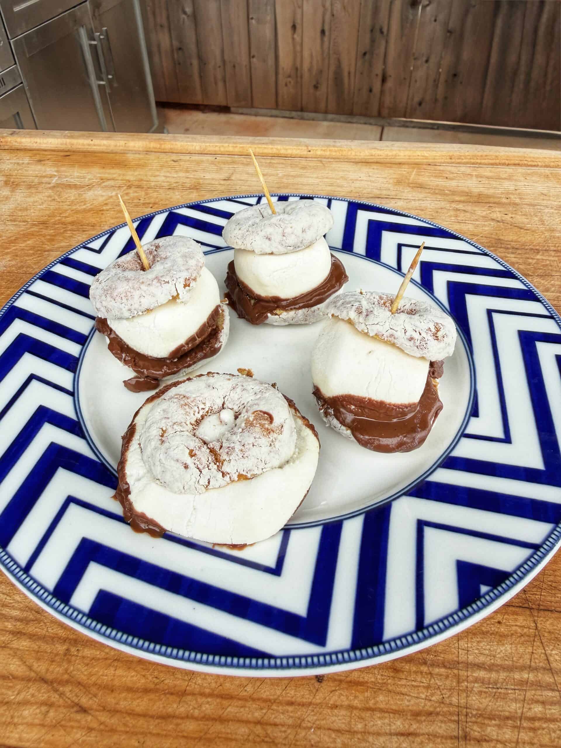 A plate with four treats made of powdered mini cinnamon rolls, chocolate spread, and large marshmallows held together with toothpicks, on a blue-and-white geometric patterned plate.
