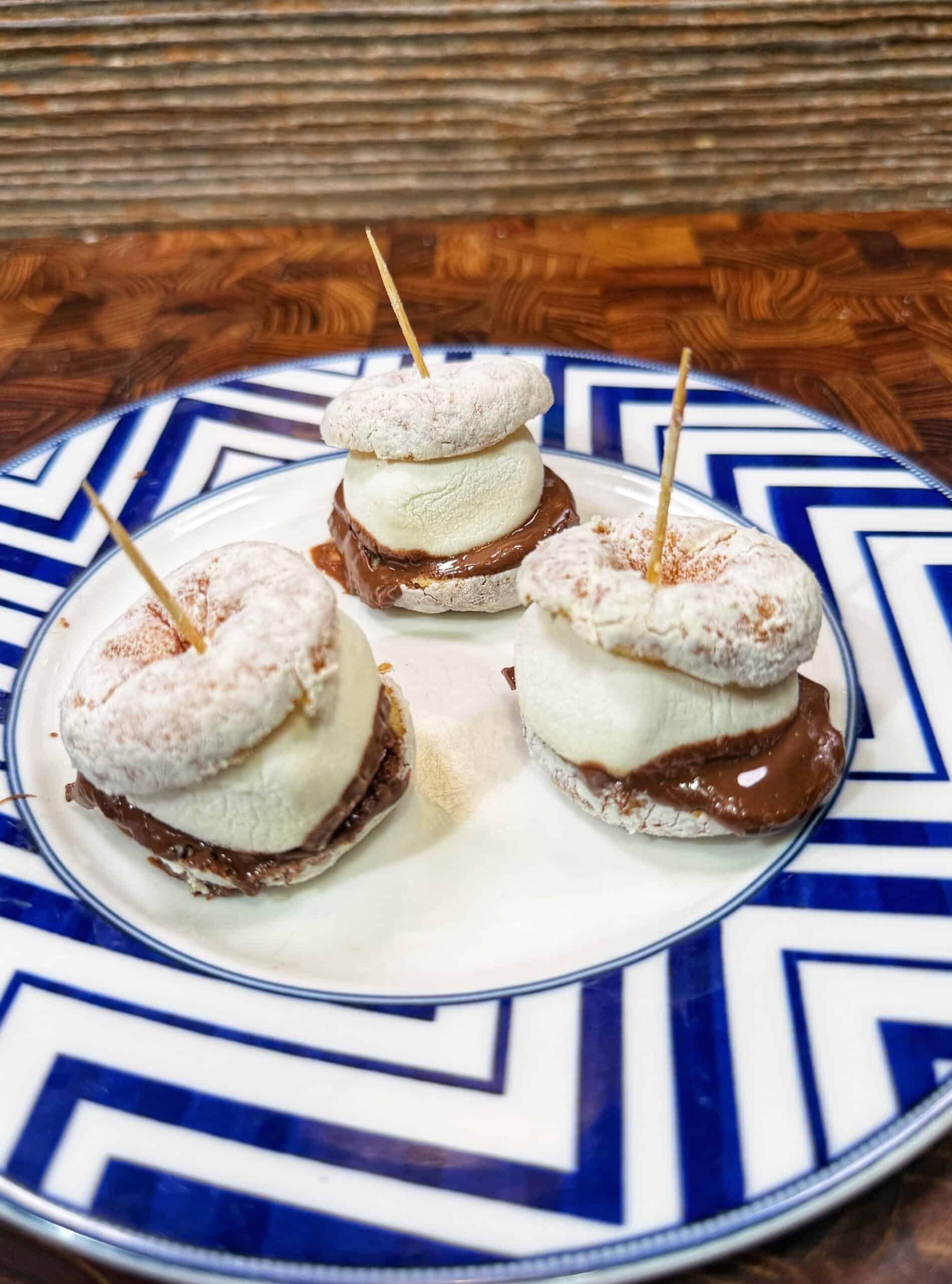 Three snack bites with powdered donuts, marshmallows, and chocolate spread are stacked and held together by toothpicks, served on a white plate with a blue geometric border.