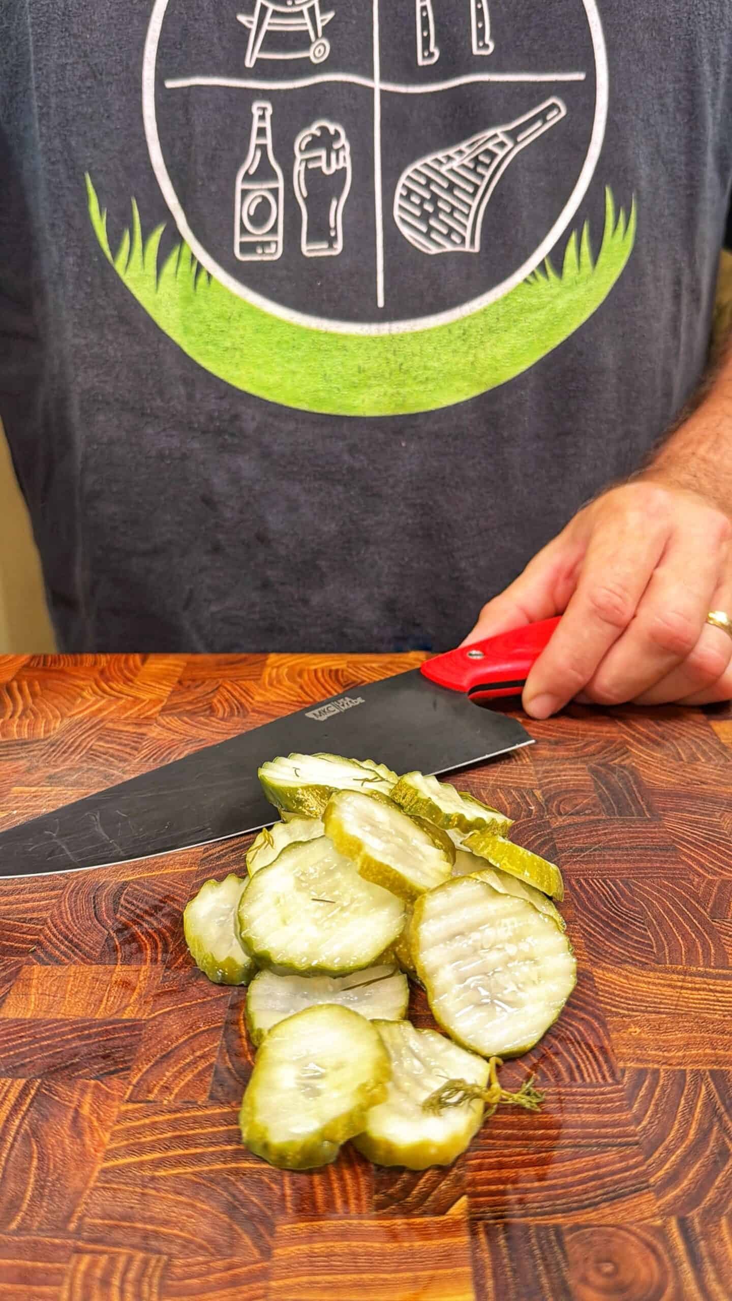A person wearing a dark shirt with graphic designs is holding a knife and slicing pickles on a wooden <a href=