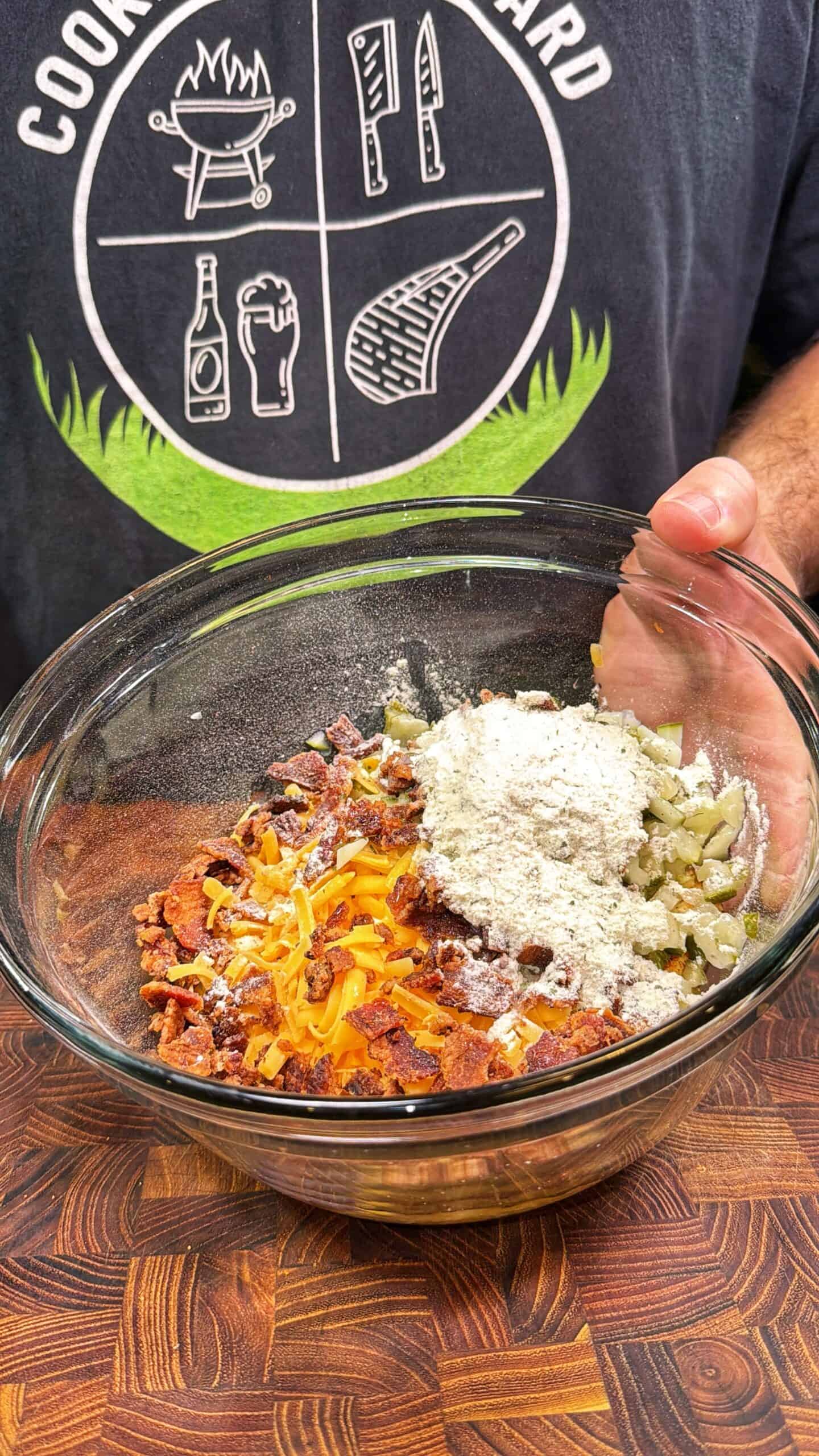 A person holding a glass bowl filled with grated cheese, bacon bits, chopped vegetables, and flour on a wooden surface. The person is wearing a shirt with cooking-themed graphics.