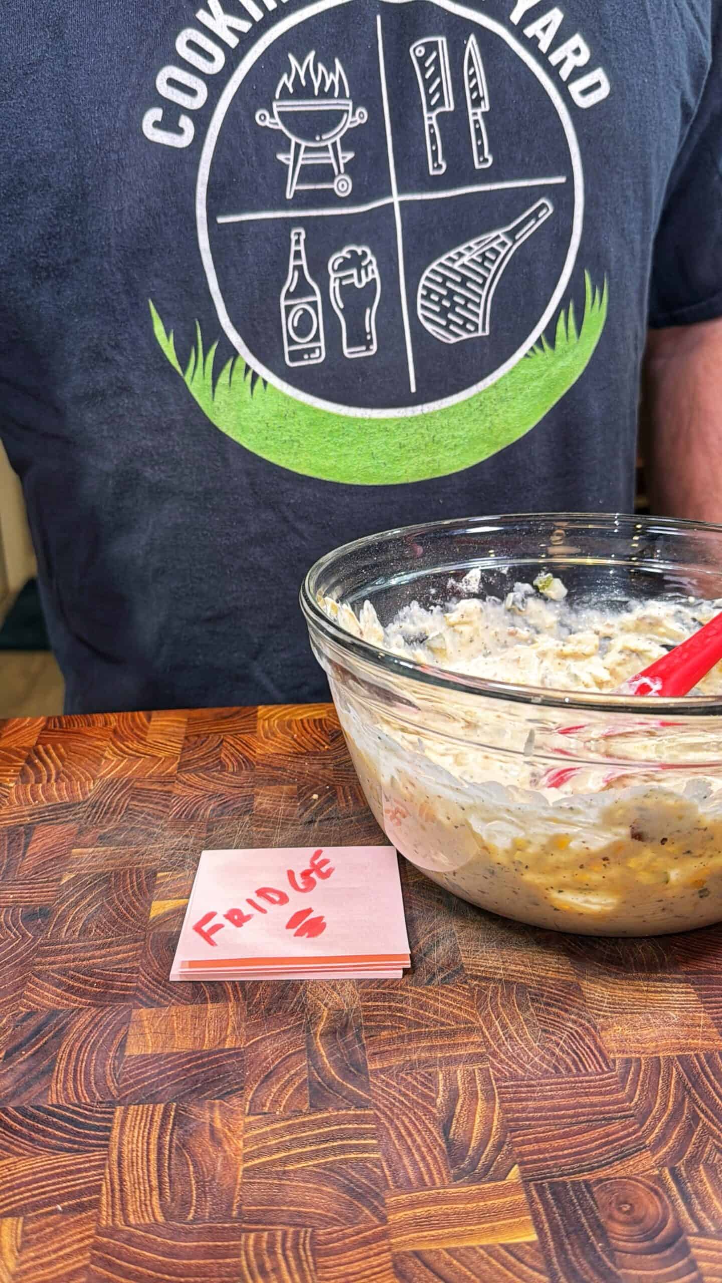 A person wearing a Cooking Wizard shirt sits behind a glass bowl filled with a creamy mixture on a wooden cutting board. In front of the bowl is a pink sticky note labeled FRIDGE with two small heart drawings.