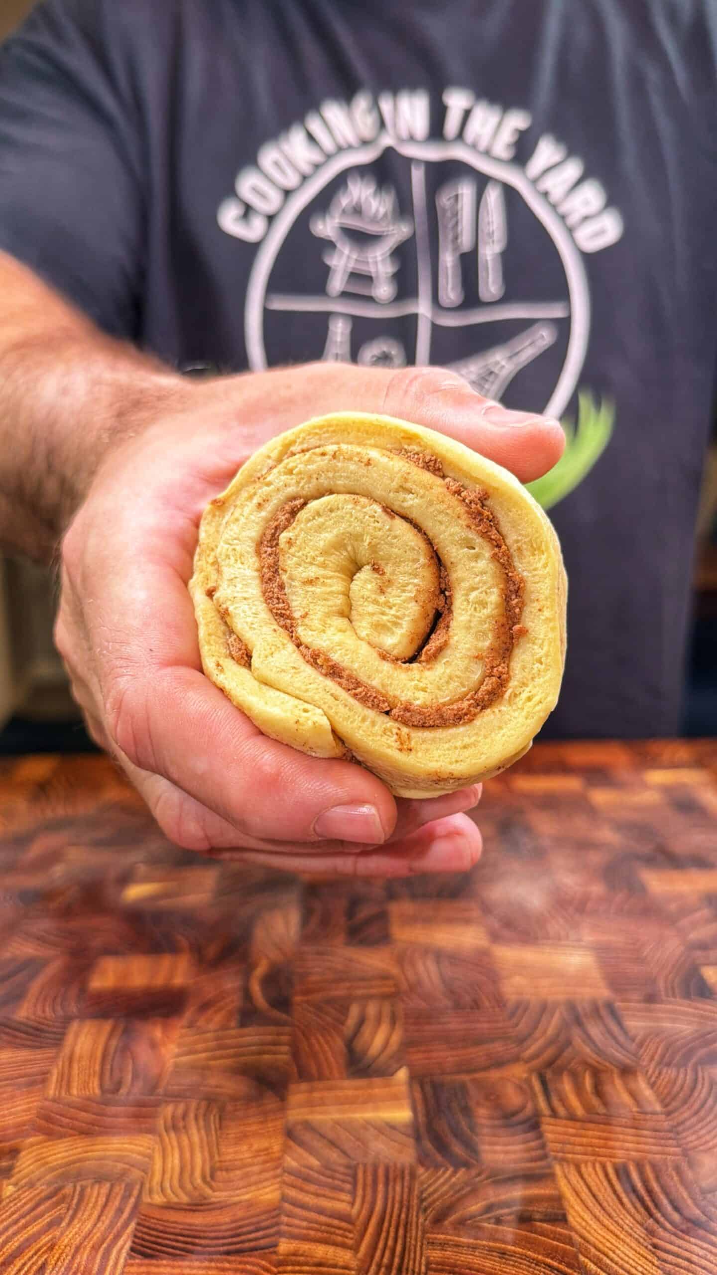 A person holds a cinnamon roll toward the camera, showing its spiral pattern. The person is wearing a dark shirt with a circular logo that says COOKING IN THE YARD. The background is a wooden surface.