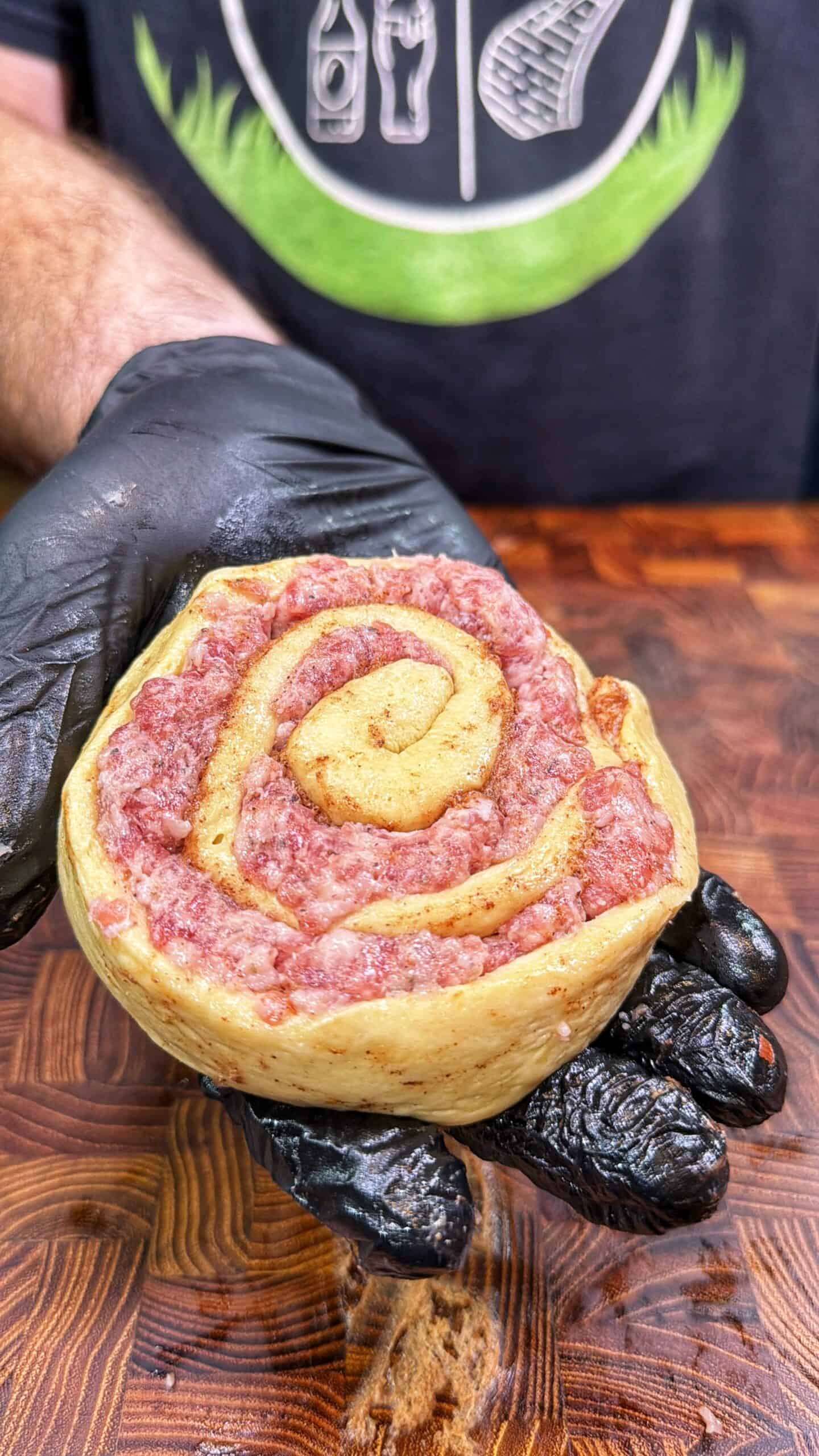 A gloved hand holds a raw meat swirl pastry that resembles a cinnamon roll, with layers of ground meat and dough visible, against a wooden surface.