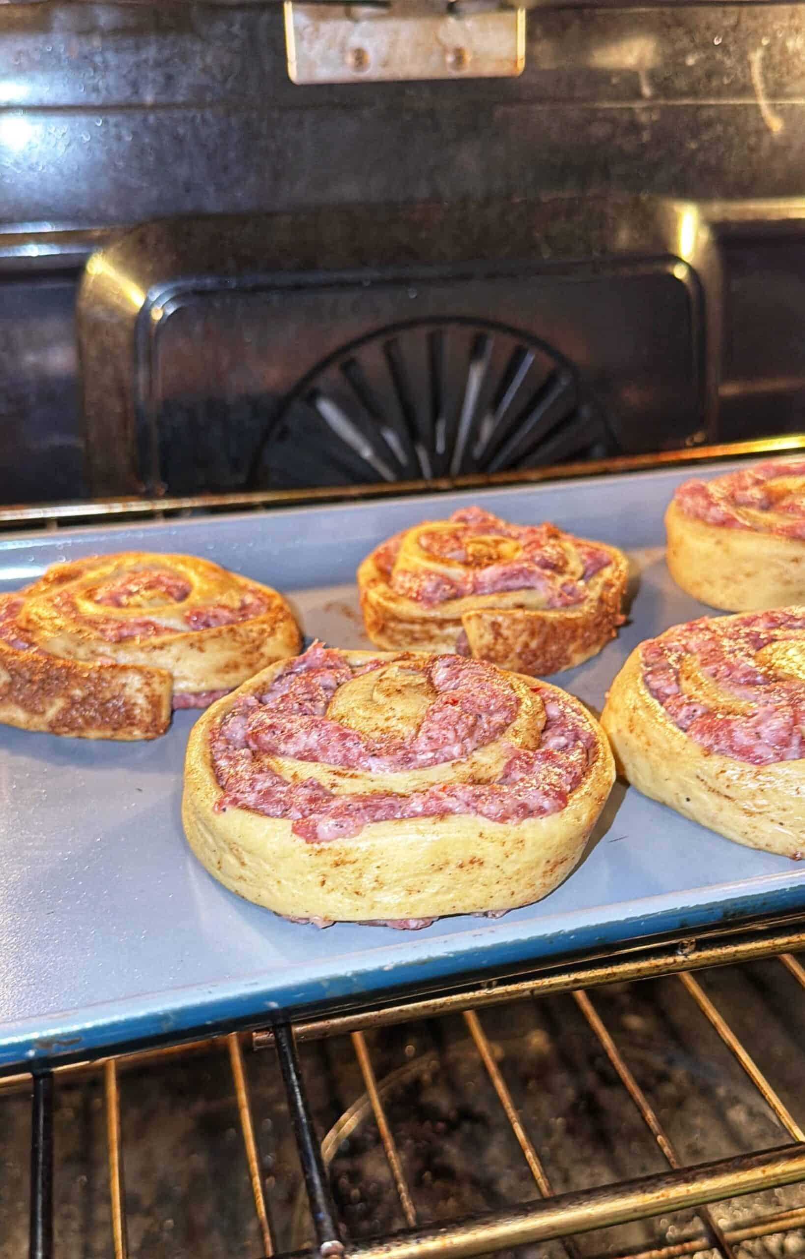 Five spiral-shaped rolls with a filling that appears to be ground meat are baking on a blue baking tray inside an oven. The rolls are golden brown and the oven is open in the background.