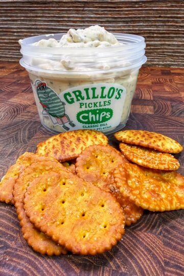 A tub of Grillos Pickles Chips Classic Dill dip sits on a wooden surface, with a pile of seasoned round and oval crackers arranged in front of it.