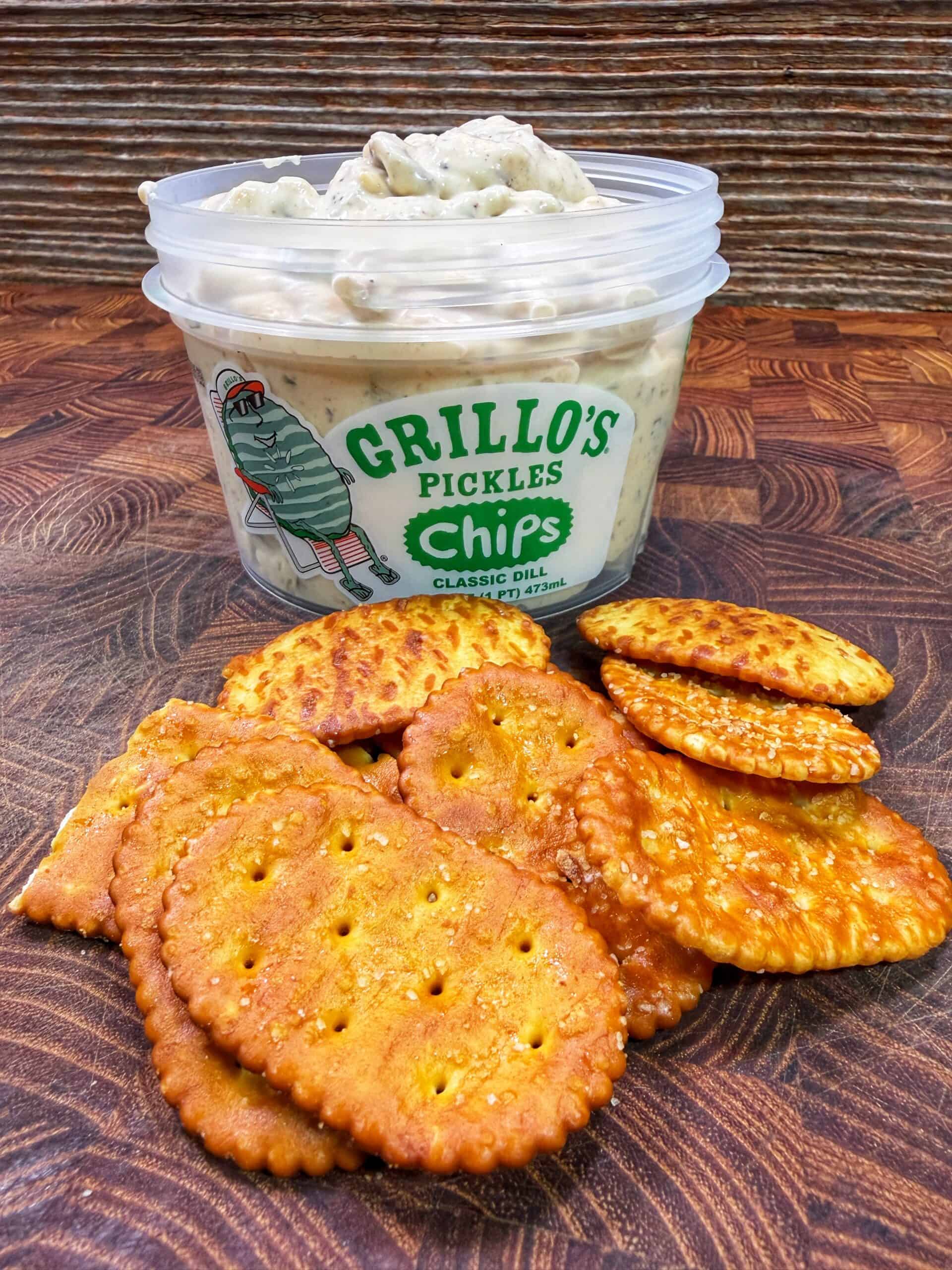A tub of Grillos Pickles Chips Classic Dill dip sits on a wooden surface, with a pile of seasoned round and oval crackers arranged in front of it.