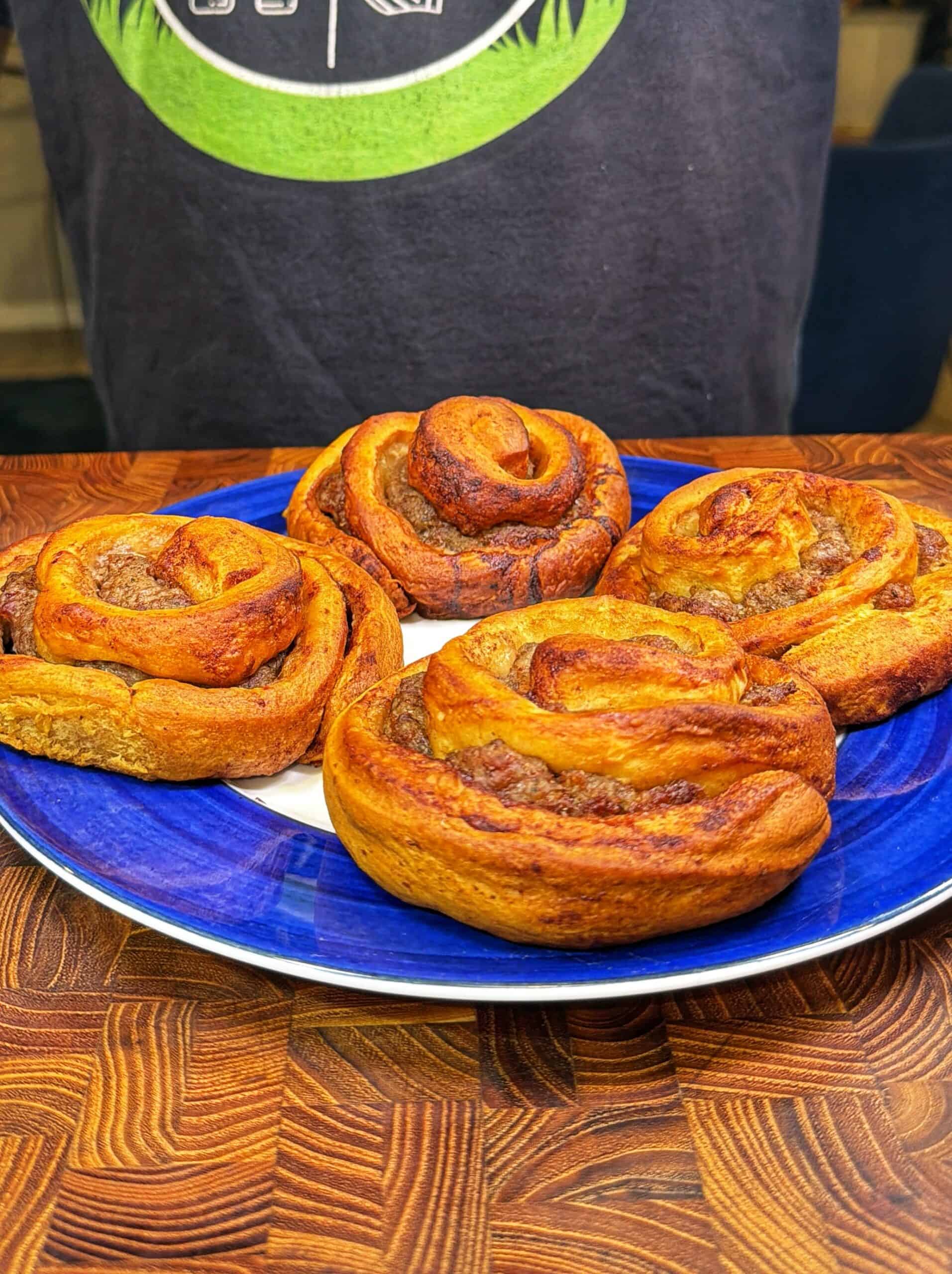 Four golden-brown cinnamon rolls arranged on a blue and white plate, placed on a wooden table, with a person in a dark shirt in the background.