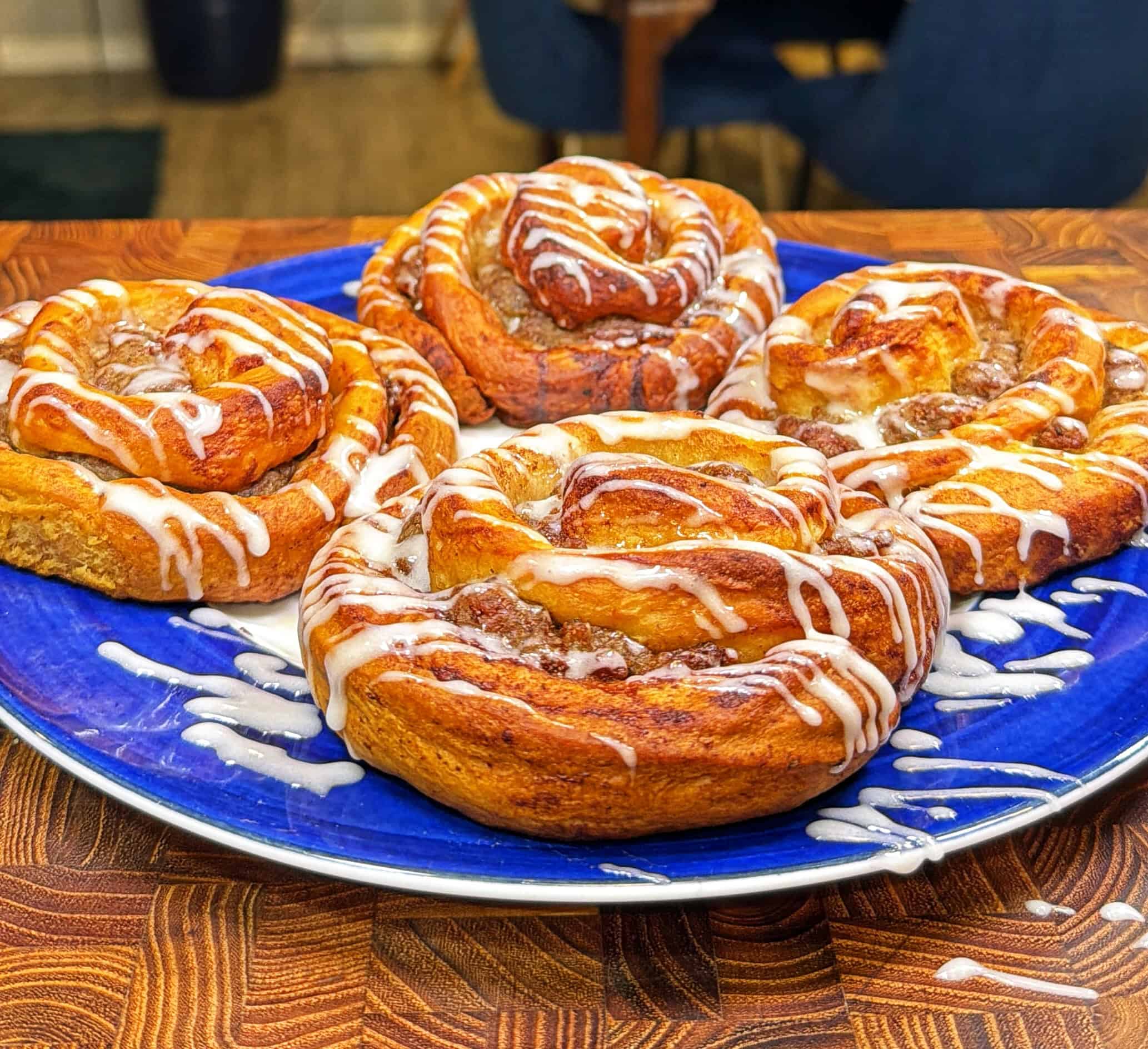 Four cinnamon rolls with white icing drizzled on top sit on a bright blue plate, placed on a wooden table. The rolls are golden brown and look freshly baked.