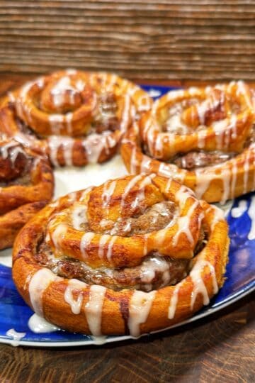 Four freshly baked cinnamon rolls topped with white icing are arranged on a blue plate, sitting on a wooden surface with a rustic background.