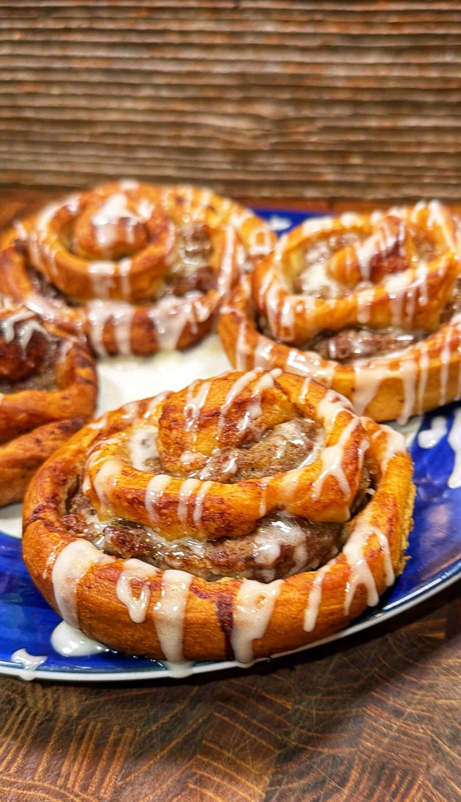 Four freshly baked cinnamon rolls topped with white icing are arranged on a blue plate, sitting on a wooden surface with a rustic background.