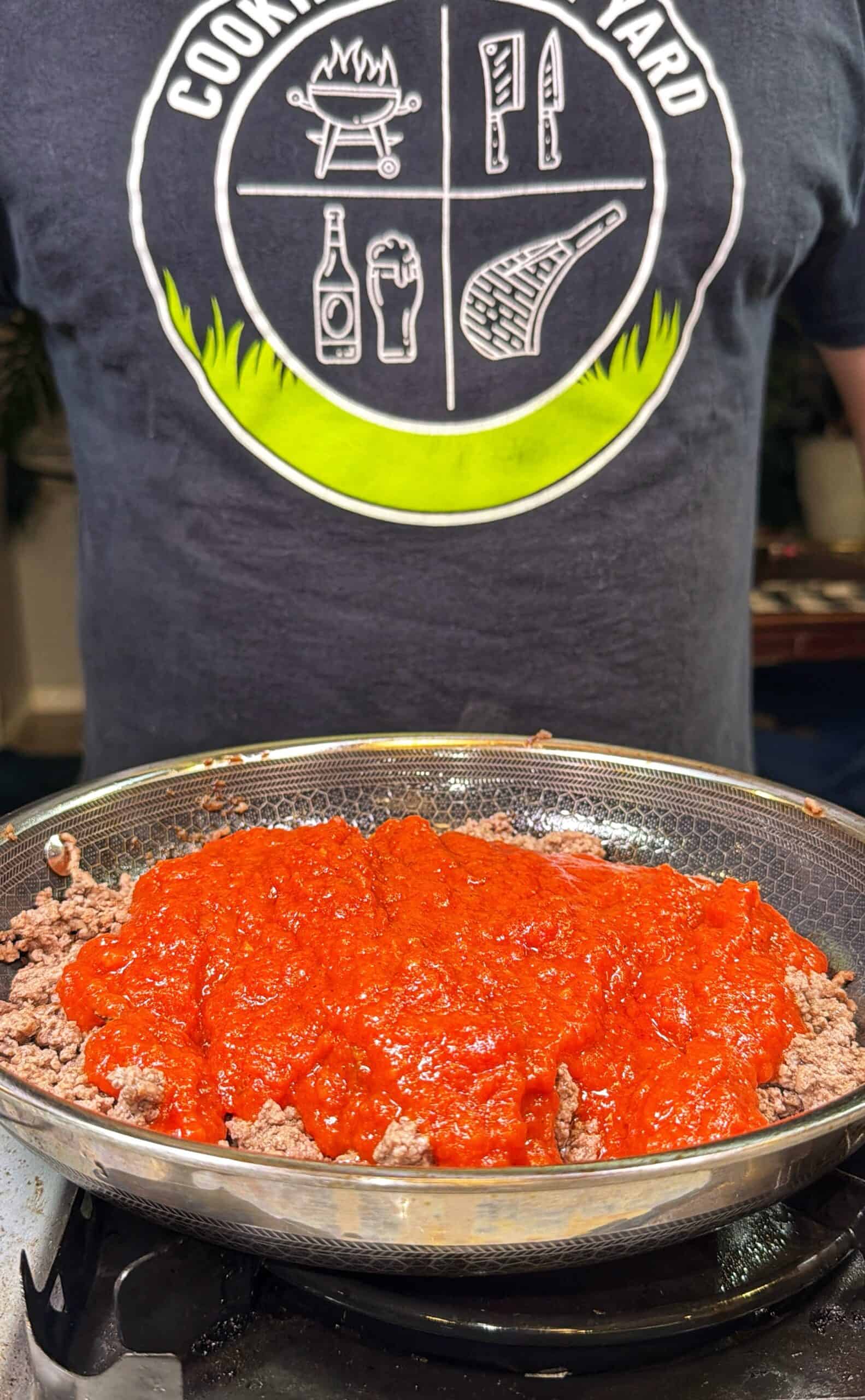 A pan of cooked ground beef topped with red tomato sauce sits on a stove, with a person wearing a Cooking Backyard T-shirt standing behind it.